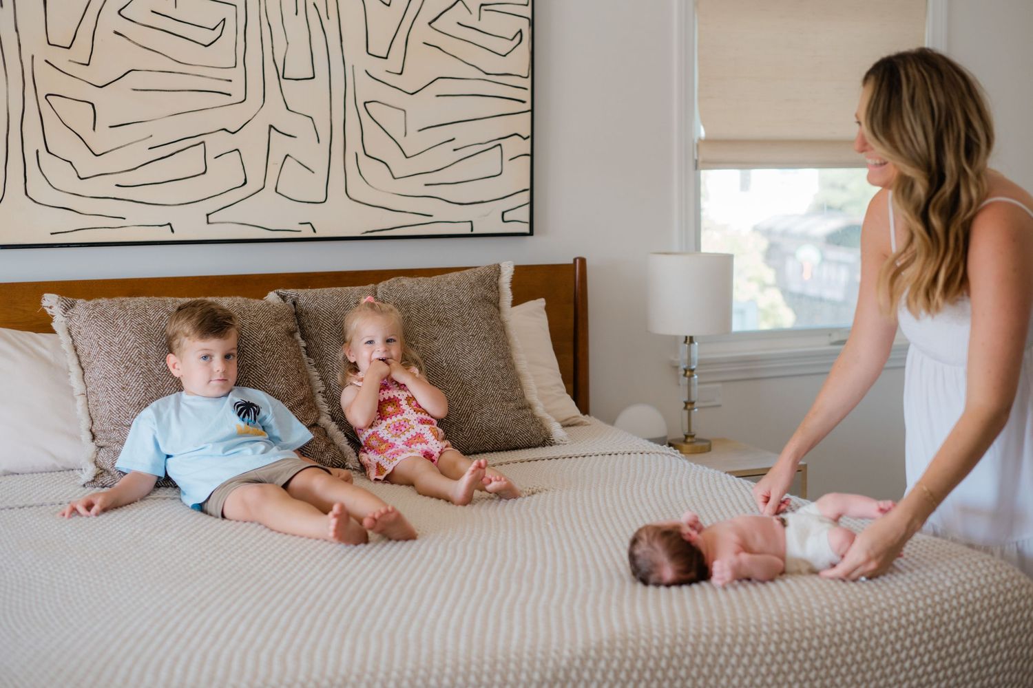 A mother tends to her newborn on a bed while two young children sit beside them in a bright bedroom.