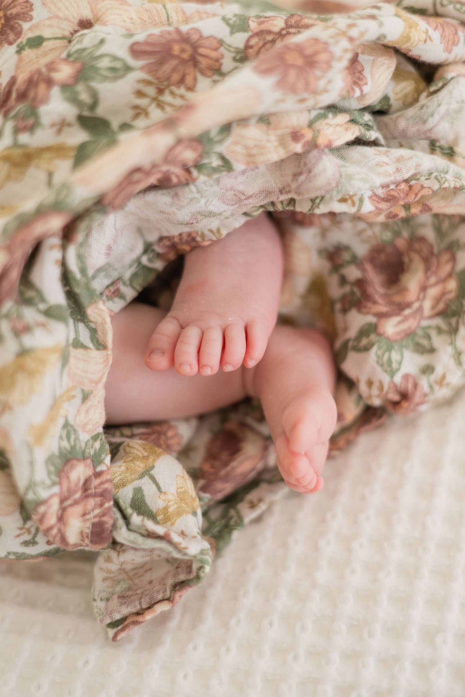 Newborn baby feet peeking out from a soft floral blanket, resting on a white textured surface.