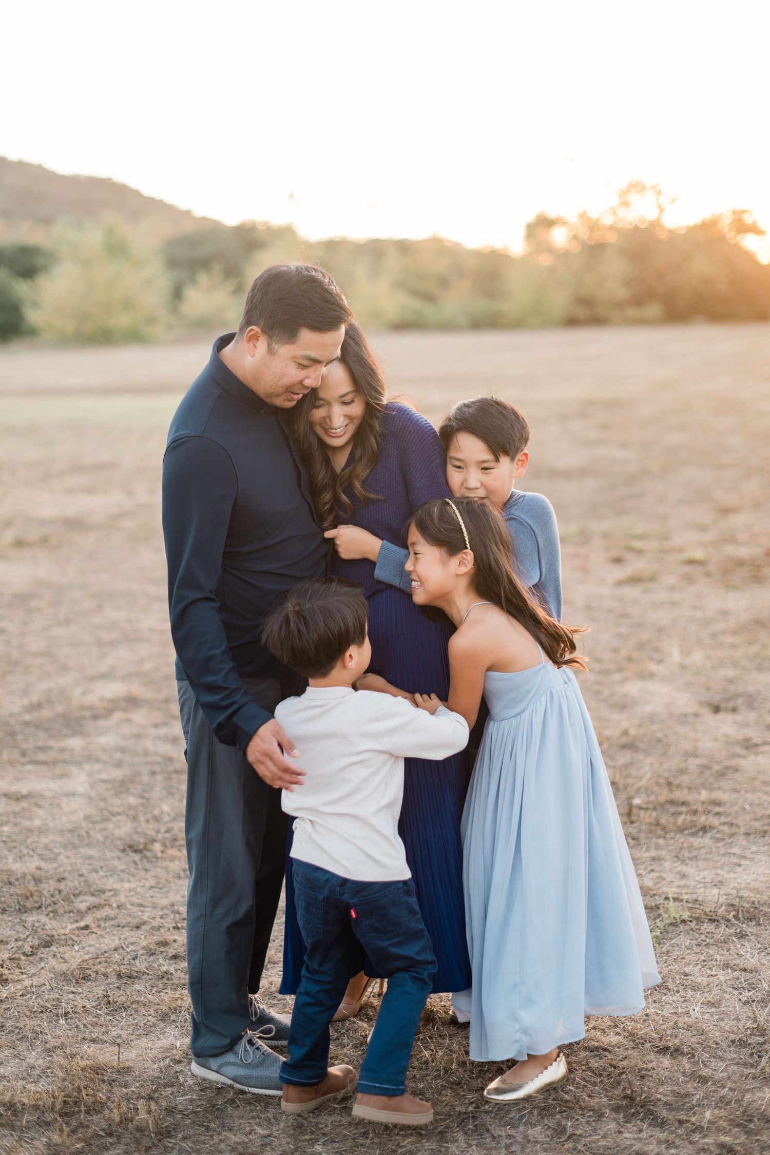 Family of five sharing a tender moment at golden hour in an open field, with children embracing their pregnant mother.