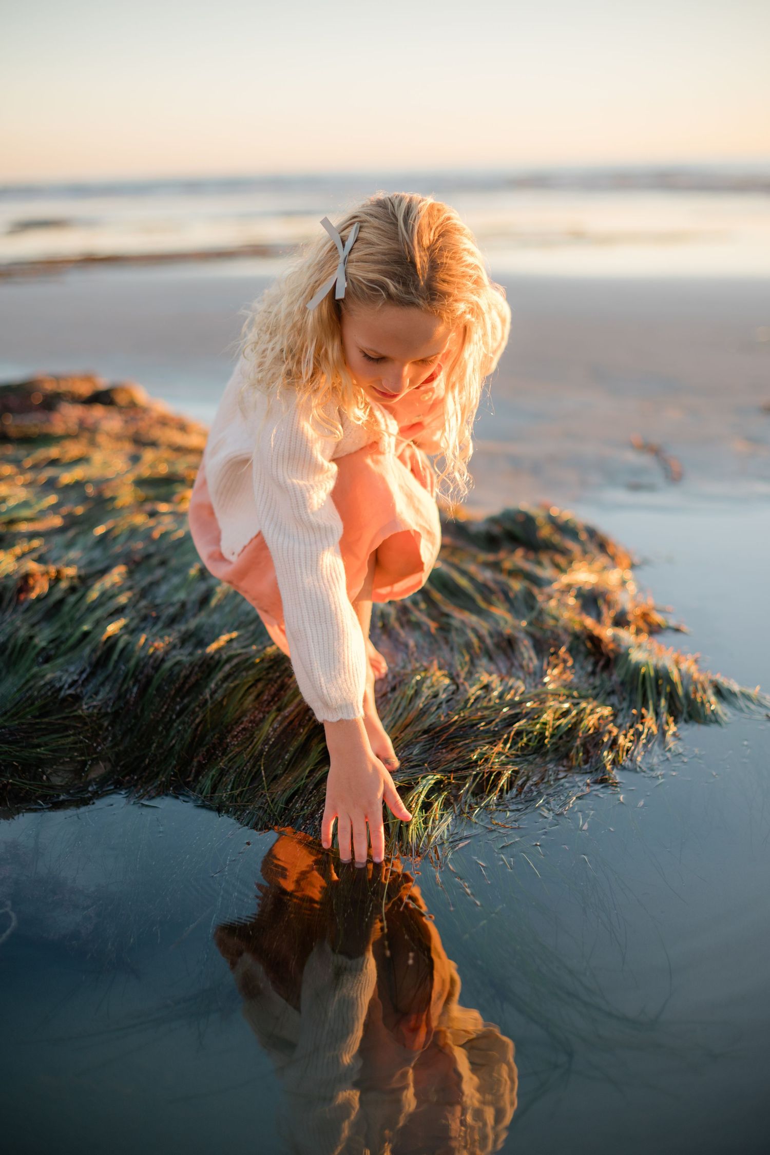 Blonde girl in pink dress crouches by tide pool at sunset beach, reaching into water.