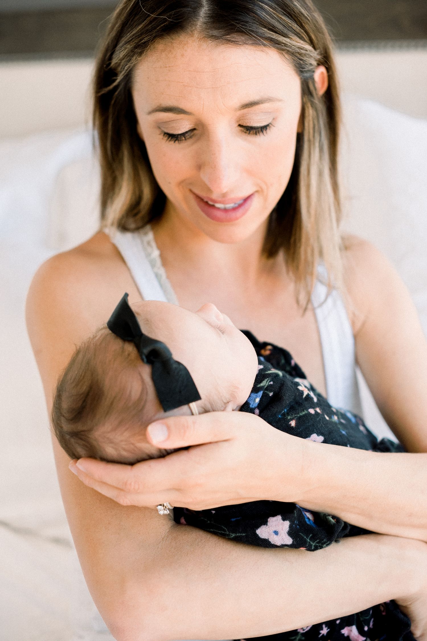 A smiling mother tenderly holds her newborn baby dressed in a dark floral outfit with a bow headband.