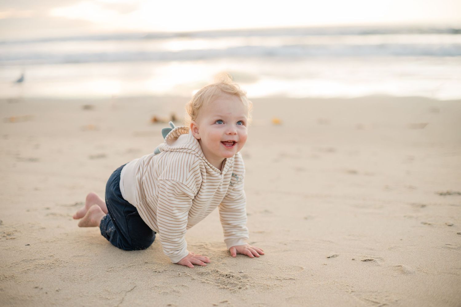 A happy baby crawls on a sandy beach near the ocean waves, wearing a striped shirt and dark shorts.
