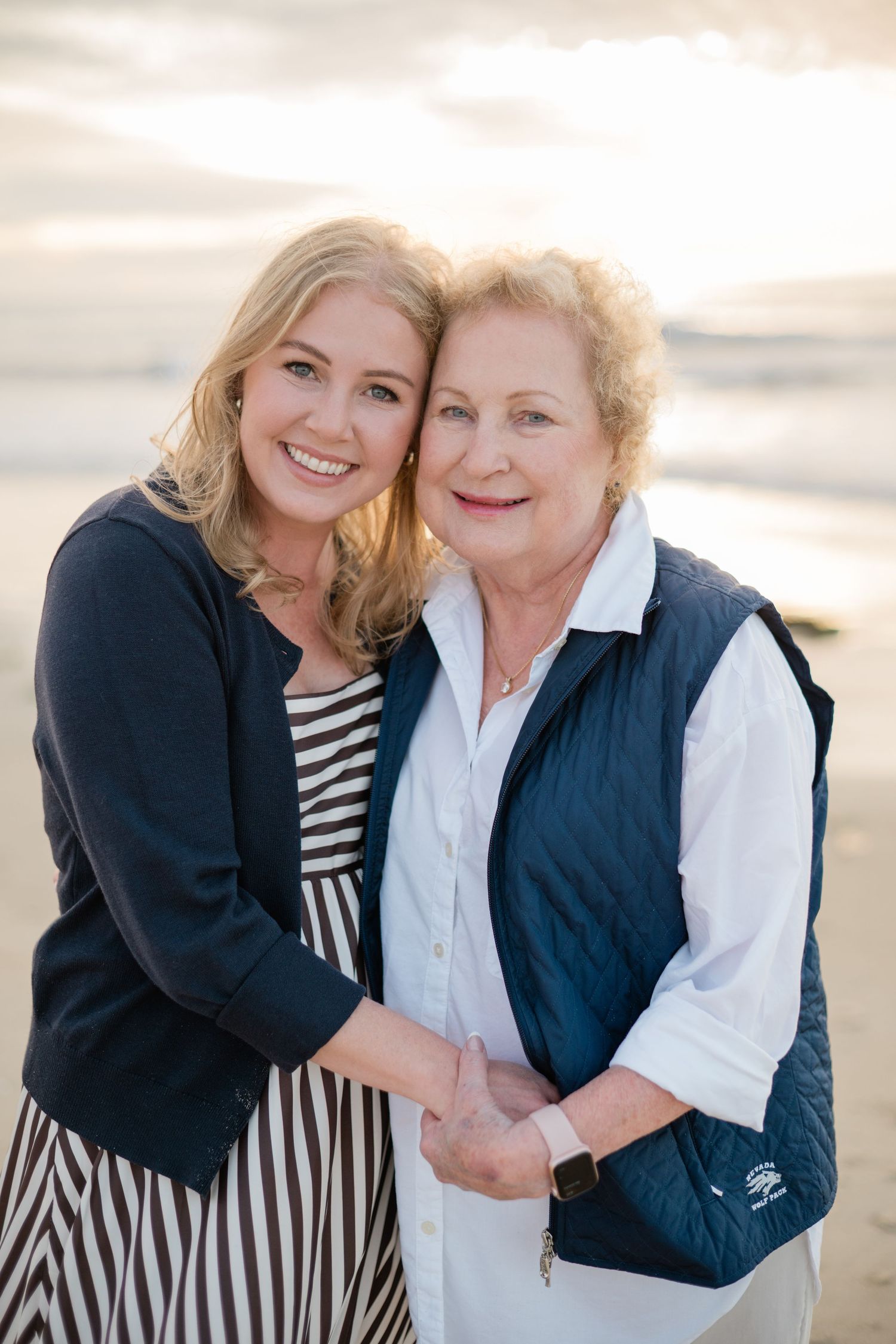 Two smiling women, likely mother and daughter, embrace on a beach at sunset wearing navy and white outfits.