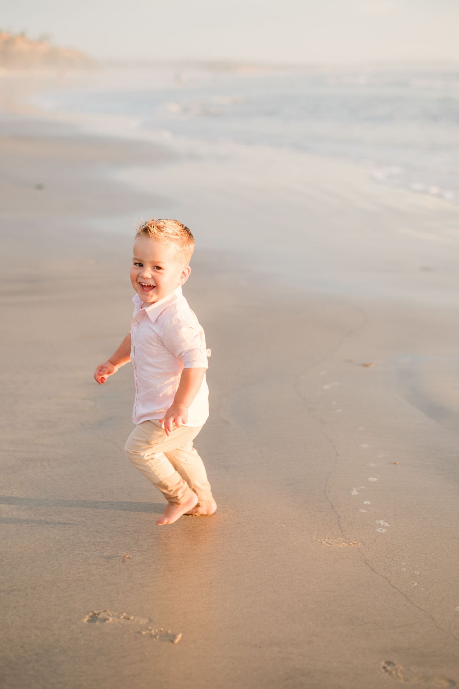 A joyful toddler boy runs barefoot on a sunny beach, laughing with waves and wet sand behind him.