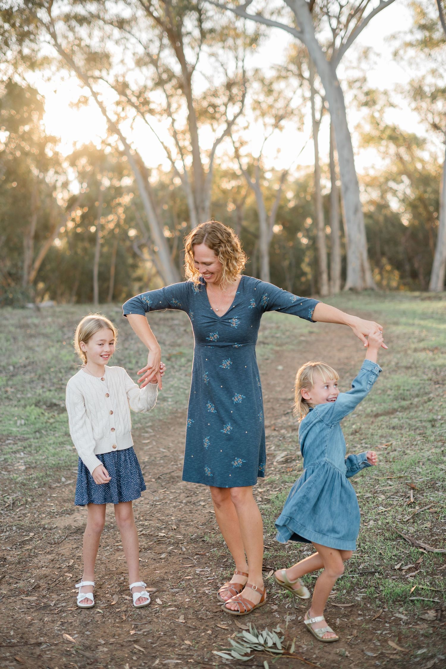 A mother dances outdoors with her two young daughters in a sunlit eucalyptus forest.