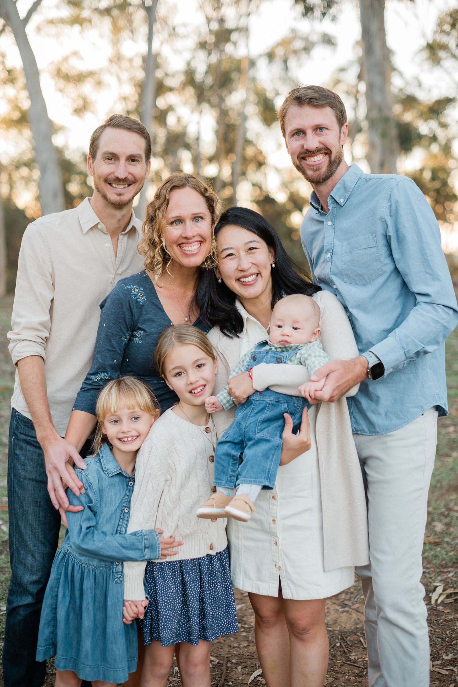 A joyful extended family group portrait with two couples, two young girls, and a baby outdoors among trees.