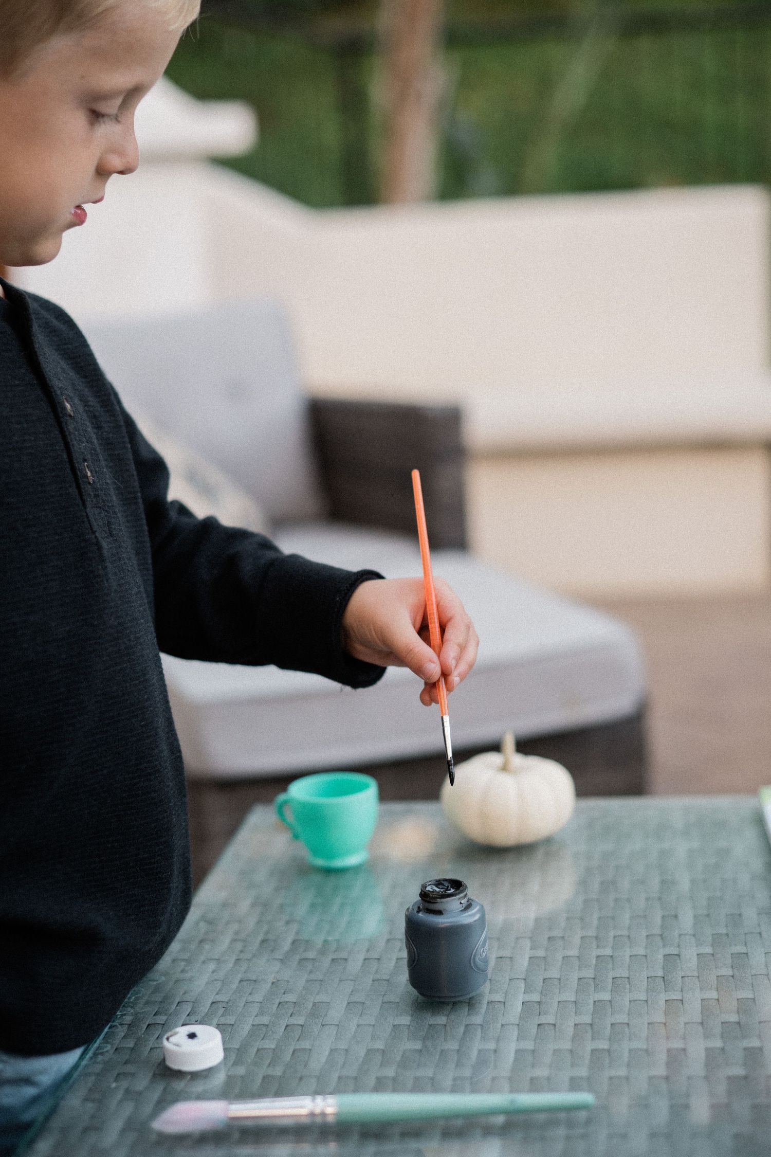 Young child painting a white pumpkin with black paint on an outdoor patio table.