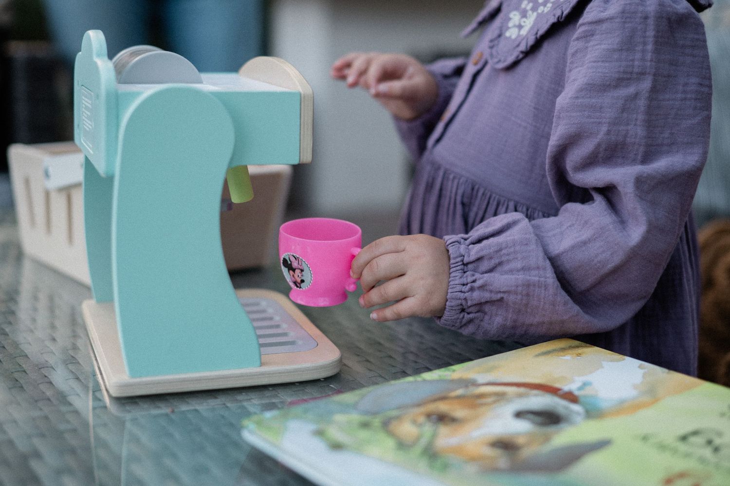 Child playing with mint green toy coffee machine and pink cup on table.