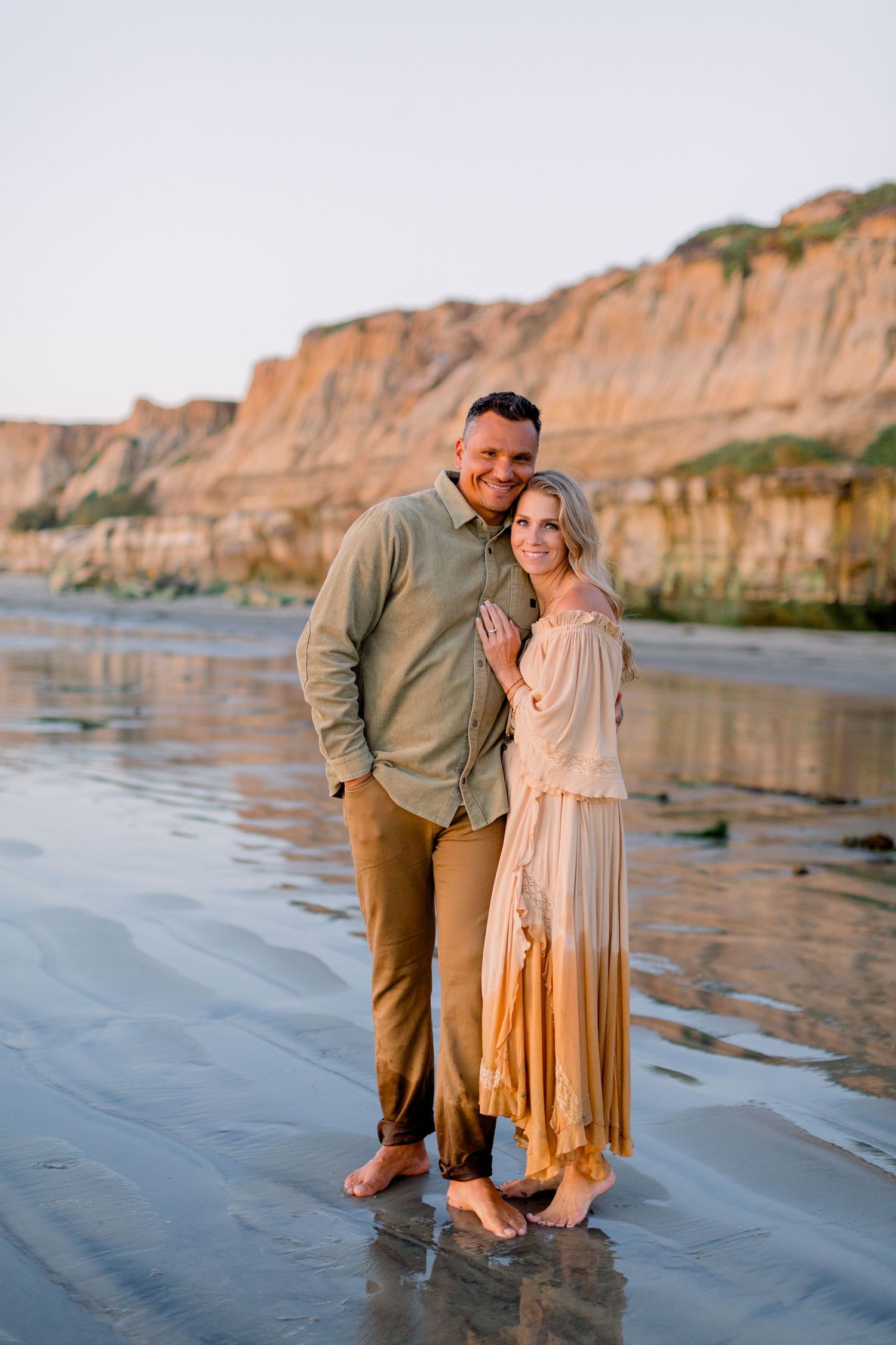 Couple standing barefoot on a reflective beach at golden hour with sandy cliffs in the background.