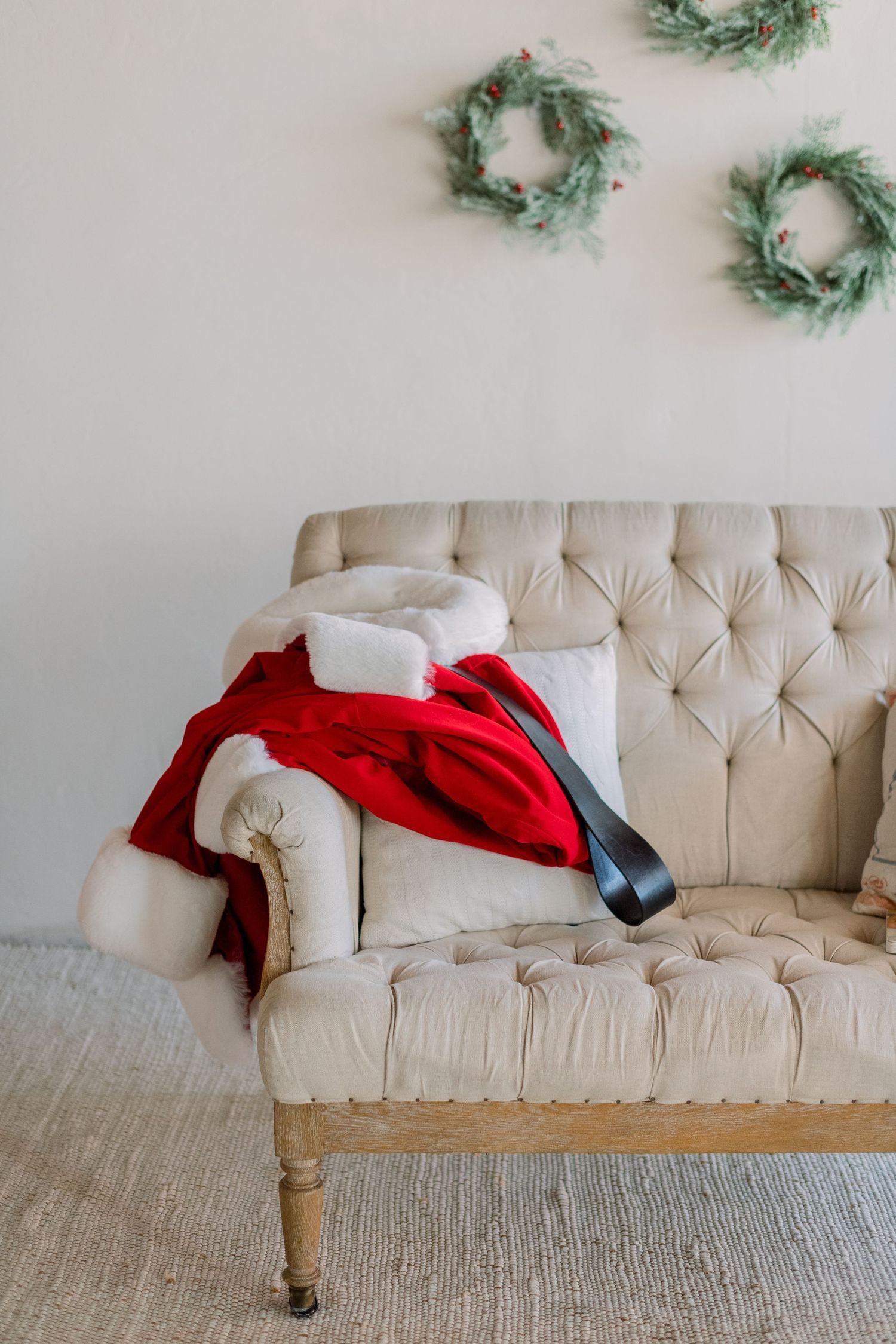 Santa Claus costume with red coat, white trim, and black belt draped over a tufted beige sofa with Christmas wreaths.