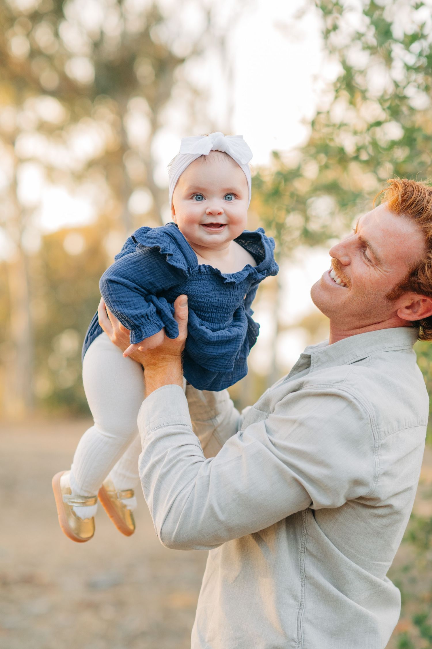 A smiling father with red hair lifts his happy baby girl outdoors, she wears a blue dress and white bow headband.