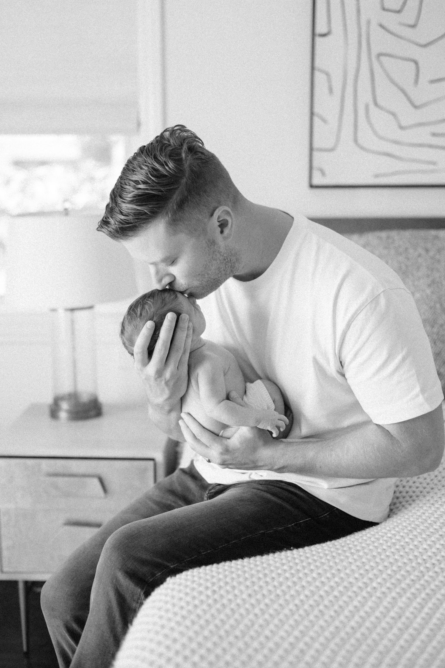 Black and white photo of a father tenderly kissing his newborn baby while seated on a bed indoors.