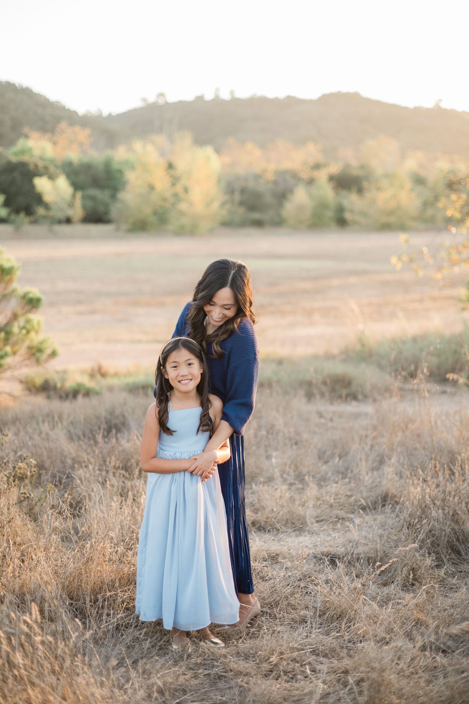 A mother and daughter in matching blue dresses pose together in a golden field at sunset.