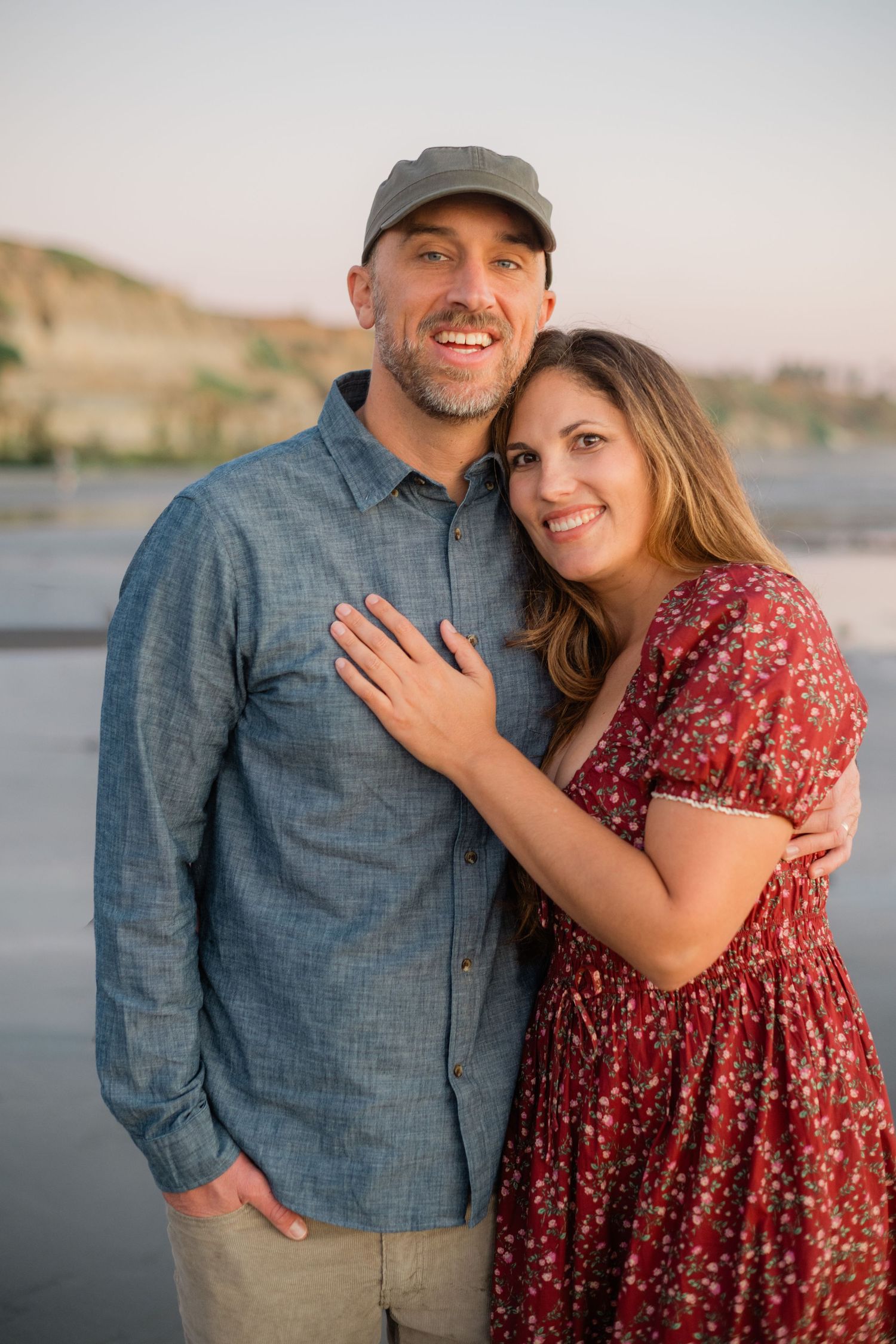Smiling couple posing together outdoors near water at sunset, woman in red floral dress.