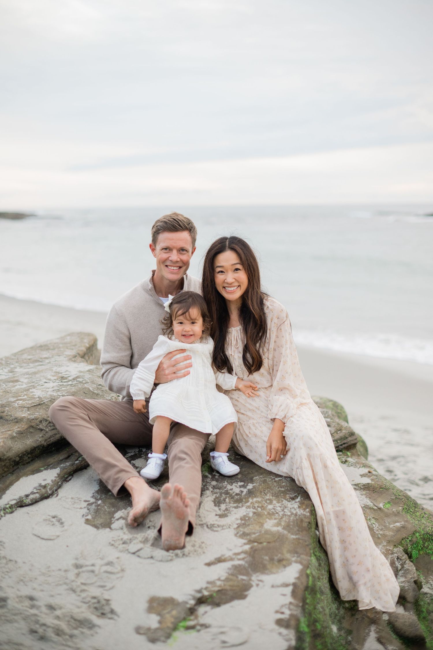 Smiling family of three posing on beach rocks with ocean waves in the background.