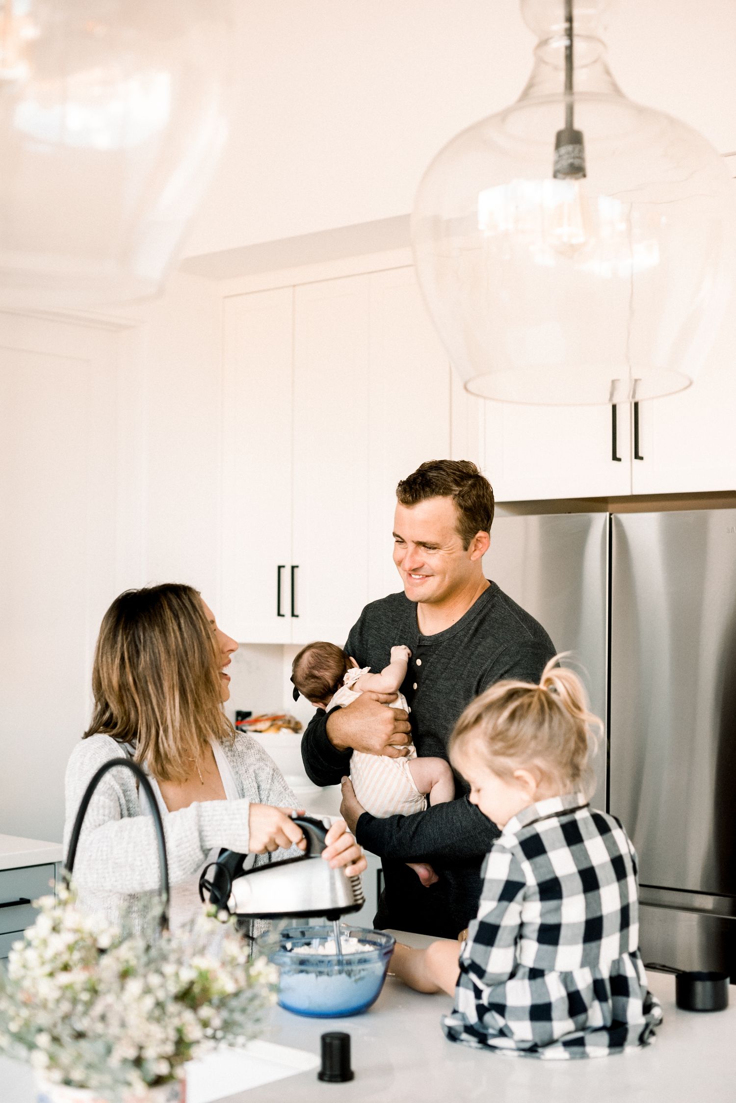 Young family cooking together in a bright white kitchen, parents smiling with two small children at the counter.