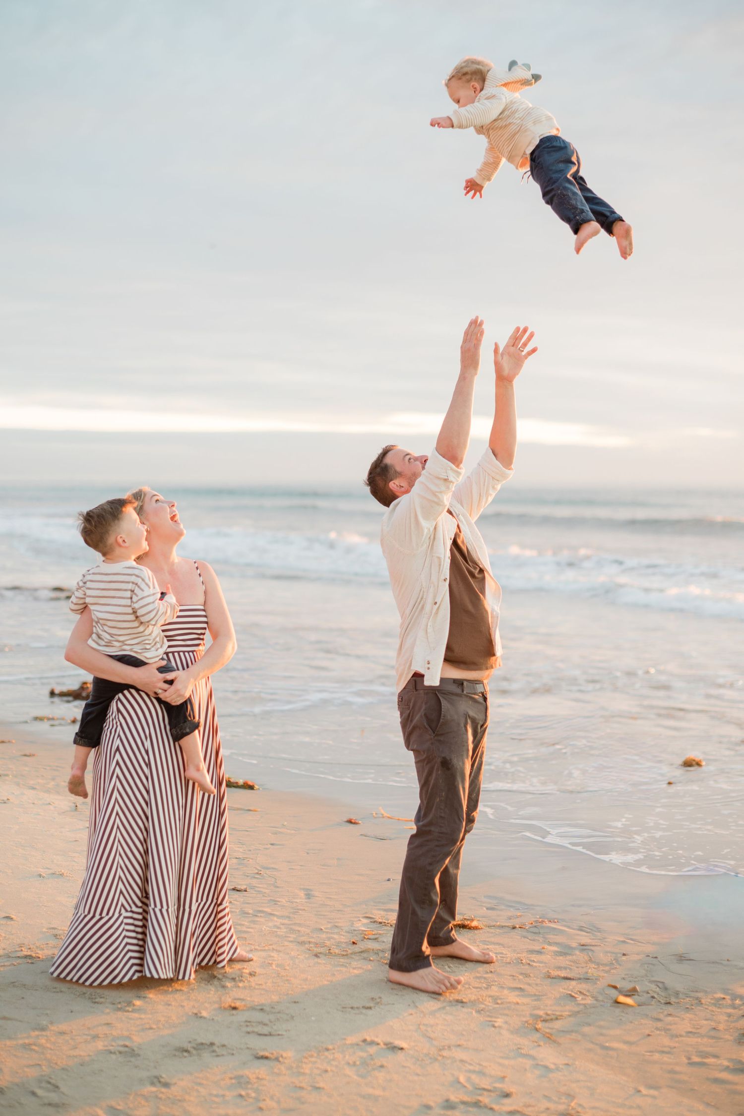 Family enjoying beach sunset, father tossing toddler in air while mother holds child on sandy shore.