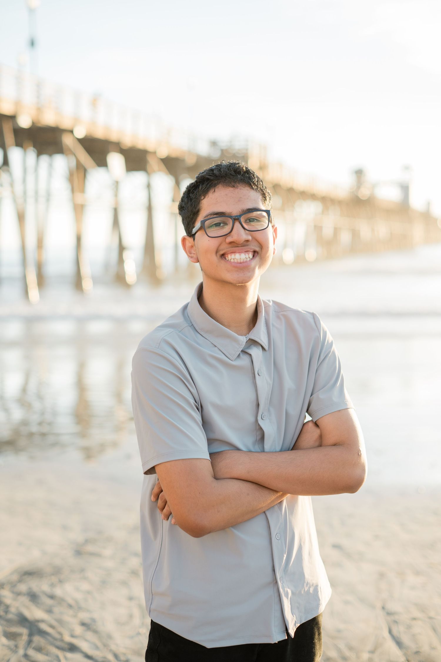 Smiling teen boy with glasses, arms crossed, standing on beach near pier in sunlight.