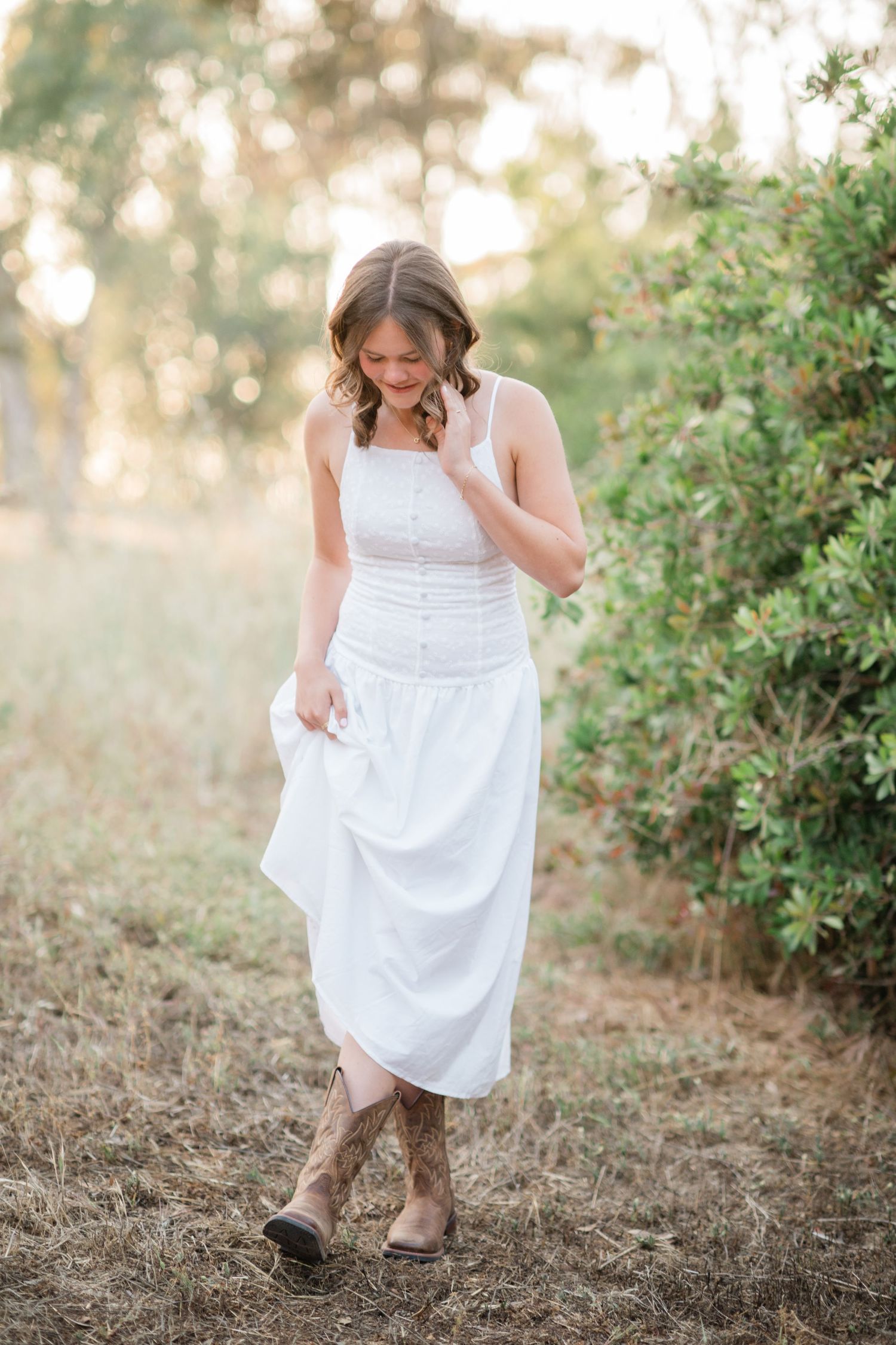 Young woman in white midi dress and cowboy boots smiles while walking outdoors in golden hour light.