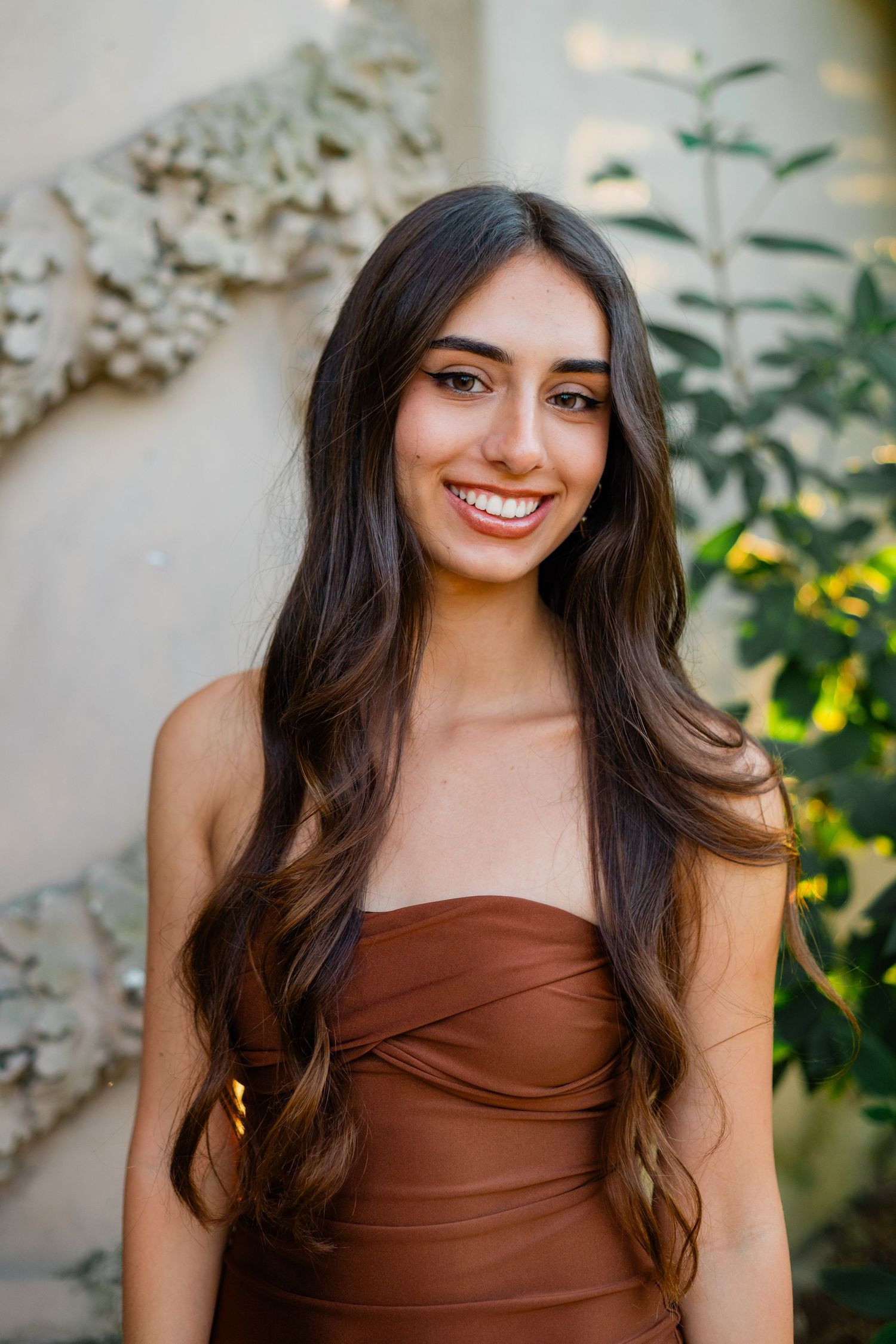 Young woman with long dark wavy hair smiles in brown strapless dress outdoors near green plants.