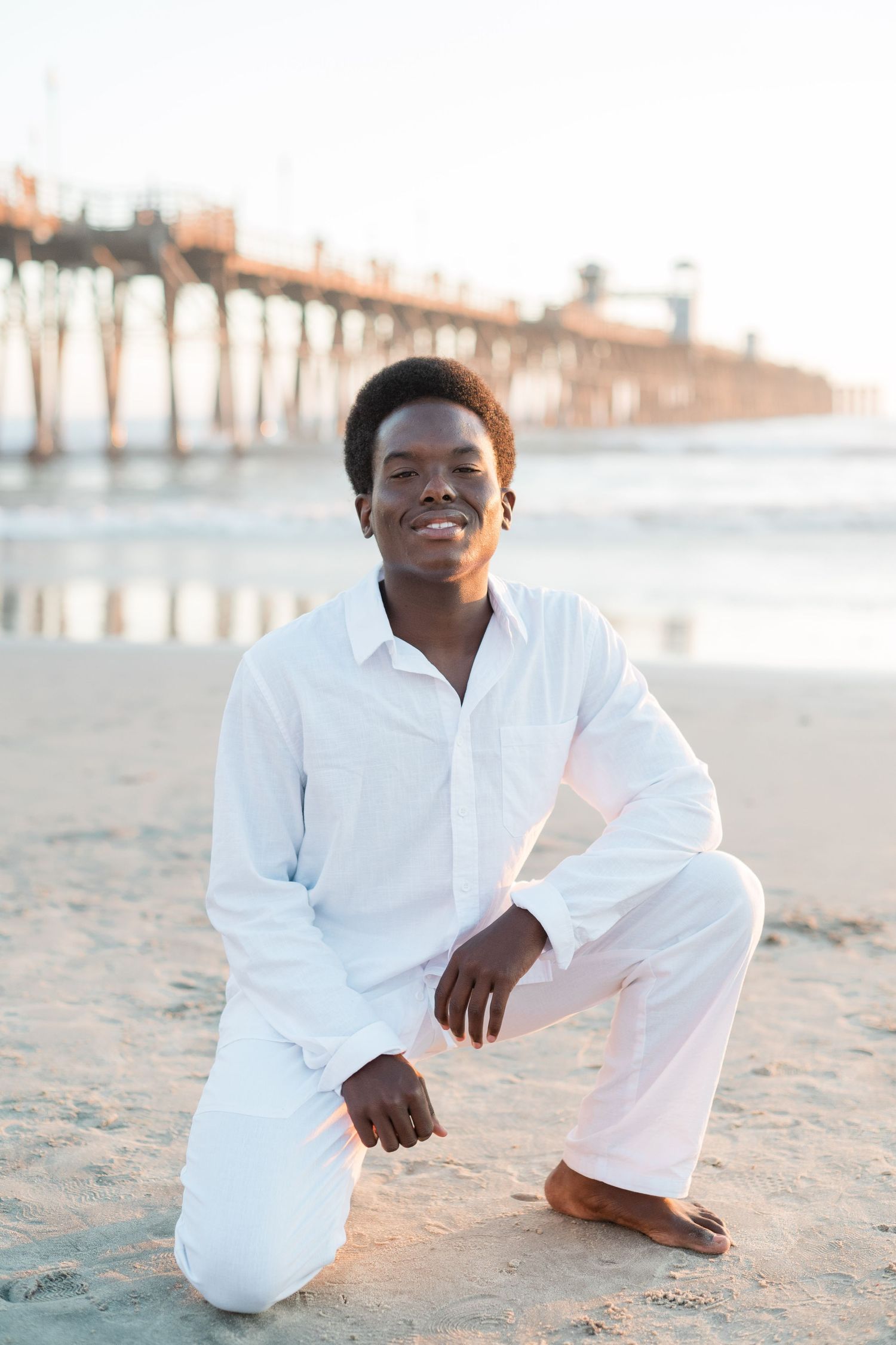 Young man in all-white outfit kneeling on sandy beach with a pier in the background at sunset.