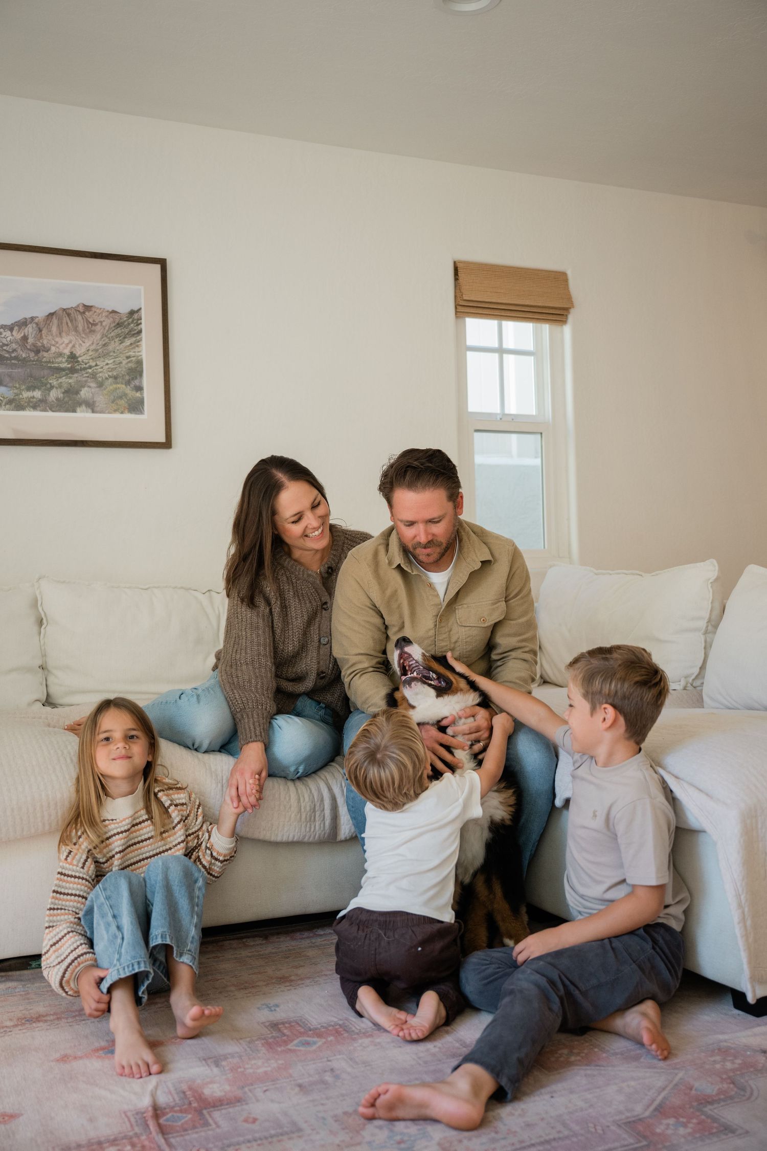 Family of four with a dog sitting together on a white sectional sofa in a cozy living room.