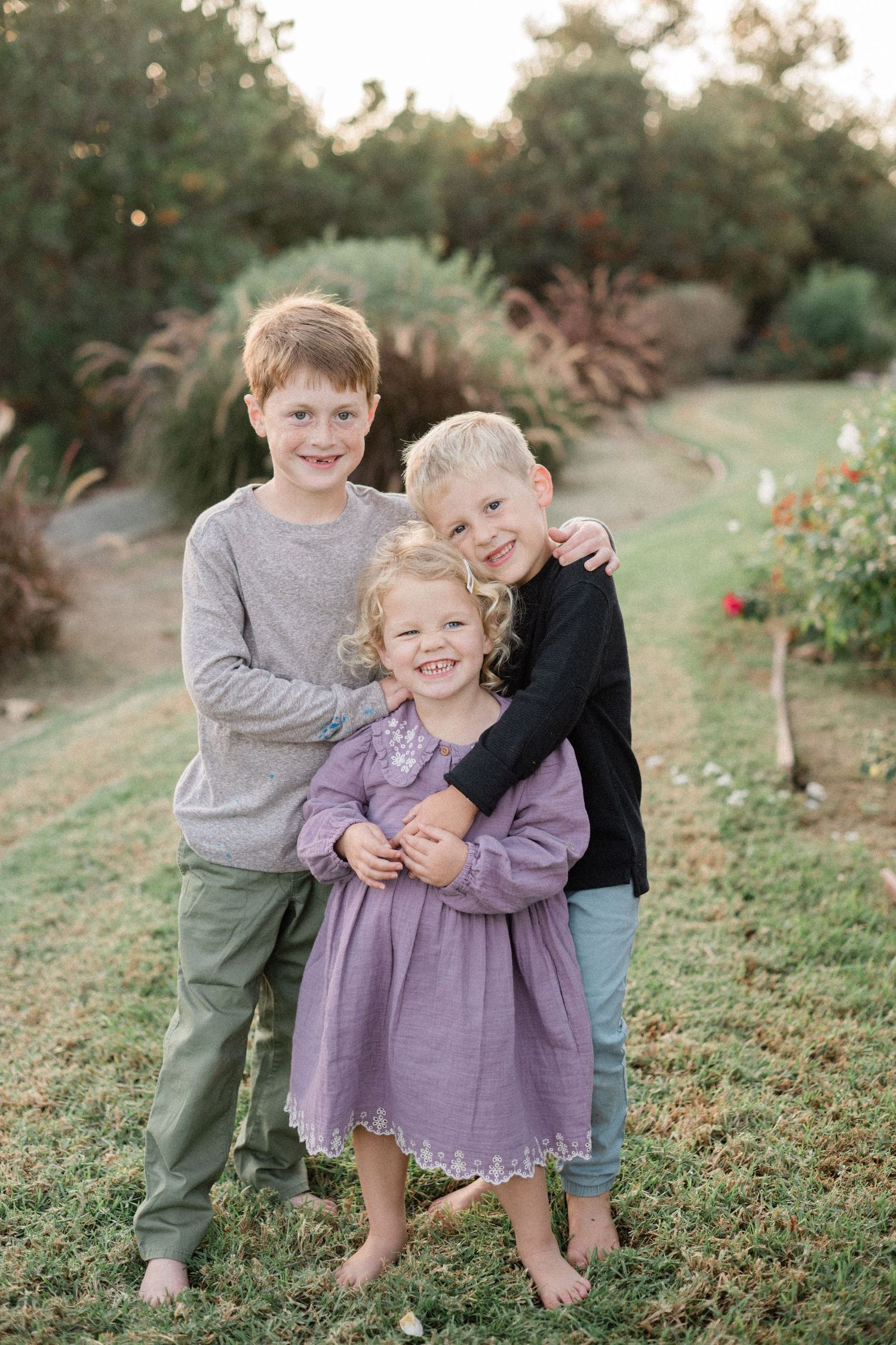 Three smiling children hugging outdoors in garden at golden hour.