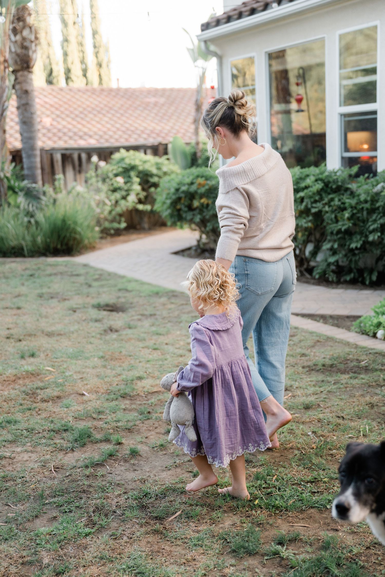 A mother and young curly-haired girl in a purple dress walk barefoot together in a lush backyard garden.