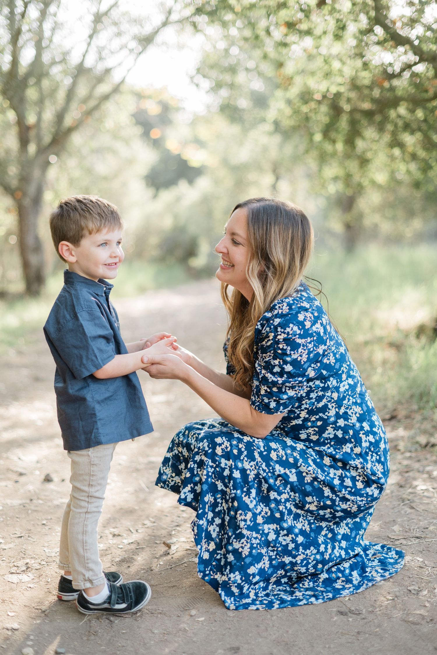 A mother in a blue floral dress kneels on a dirt path, smiling and holding hands with her young son outdoors.