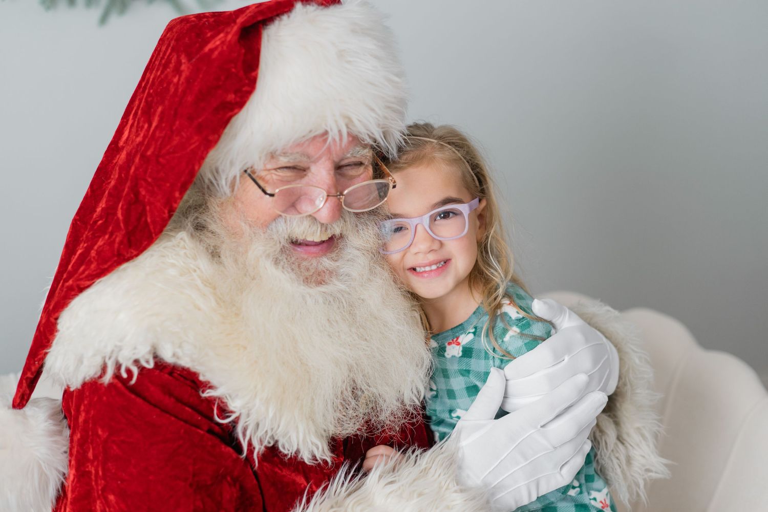 A young girl with glasses smiles while sitting with Santa Claus in his classic red suit and white beard.