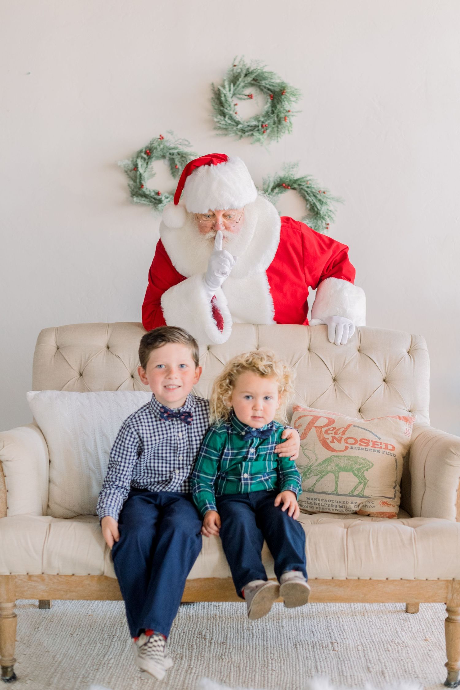 Two boys sit on a cream tufted sofa with Santa Claus making a shush gesture behind them near holiday wreaths.