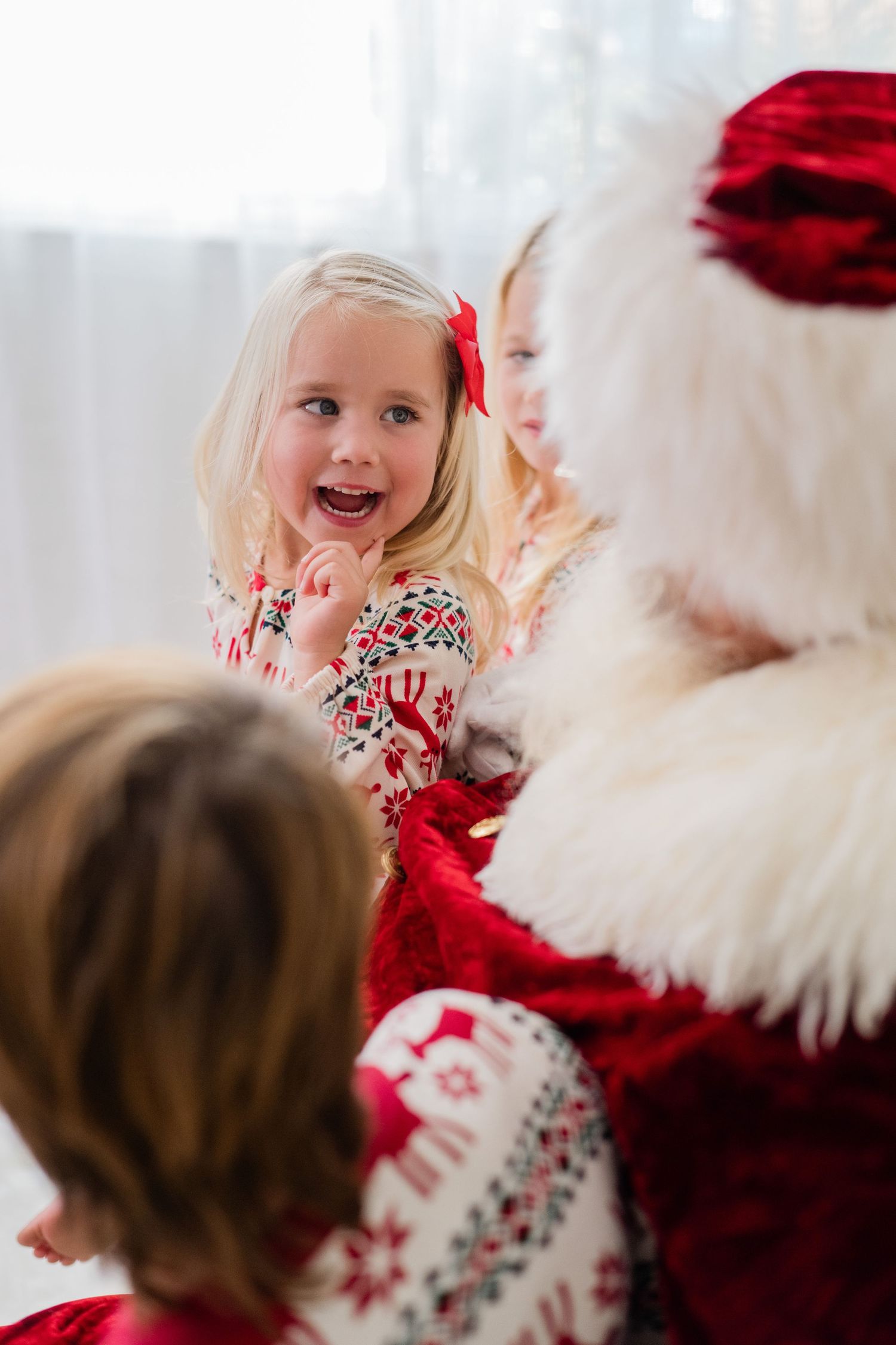 An excited blonde girl in Christmas pajamas looks up at Santa Claus with a joyful expression during a holiday visit.