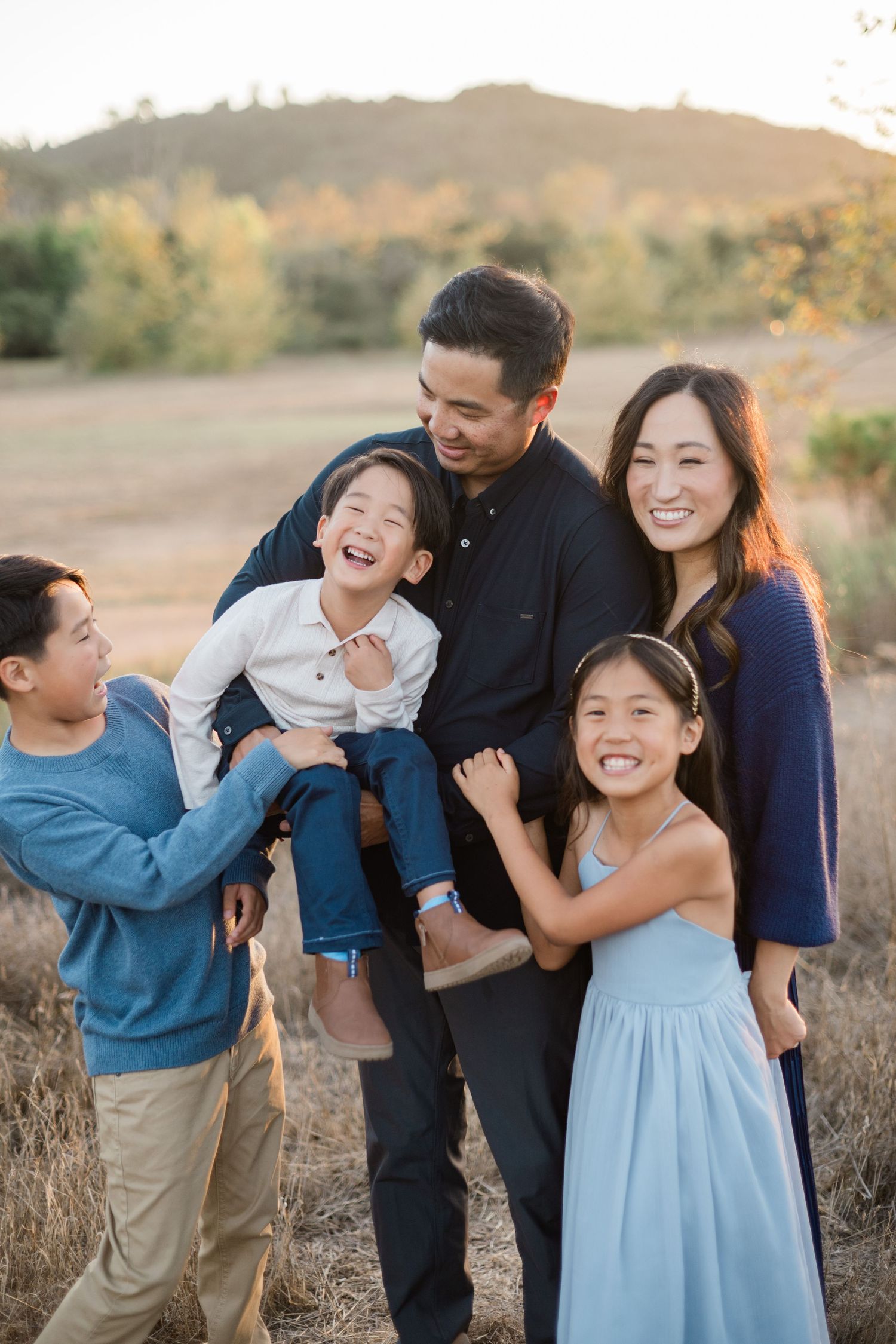A joyful Asian family of five laughs together in a sunlit field with rolling hills in the background.