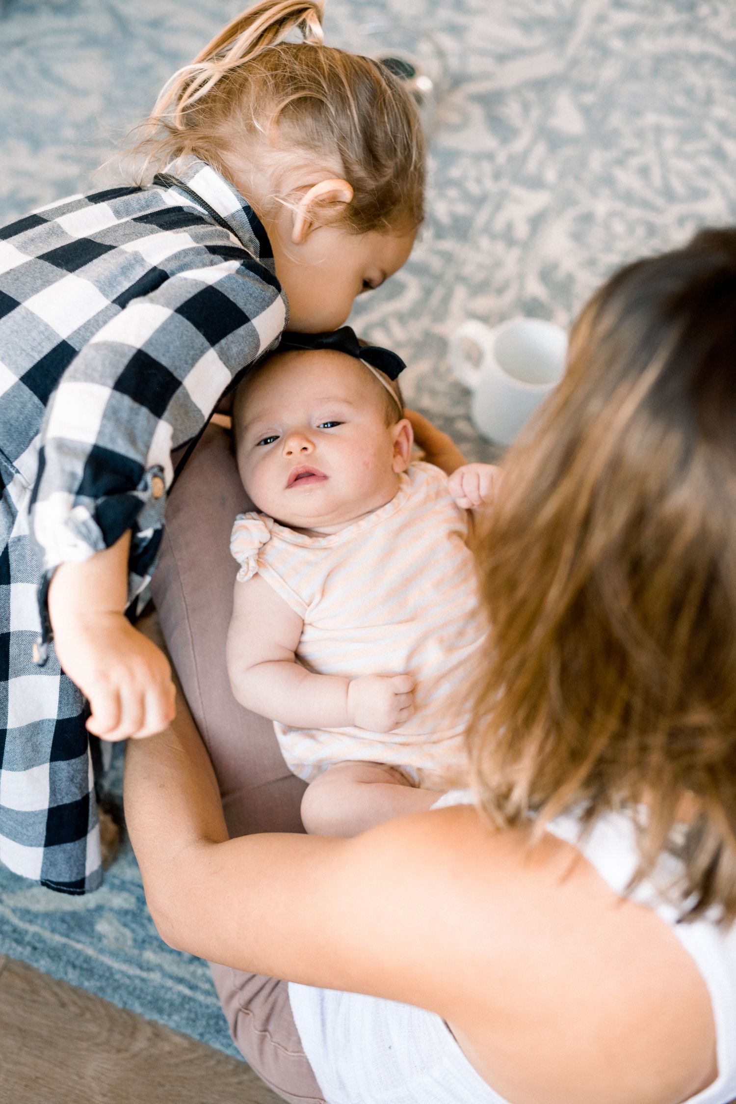A mother holds a newborn baby while a young toddler in a plaid shirt leans in to give the infant a gentle kiss.