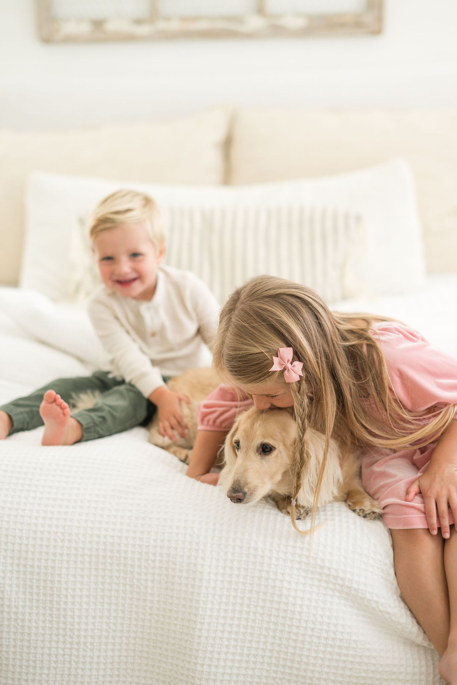 A young girl in pink hugs a golden retriever on a white bed while a laughing toddler boy sits nearby.