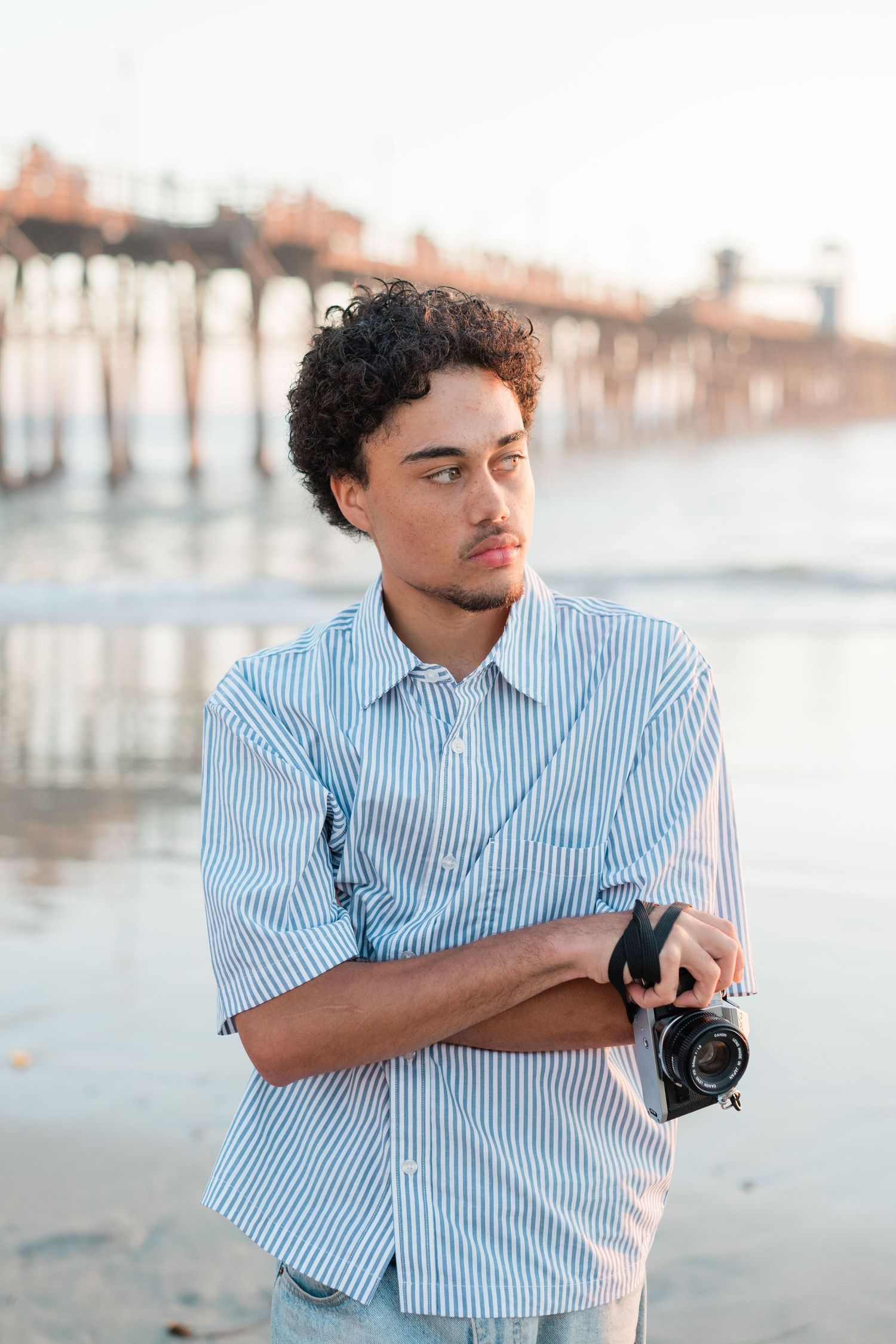 Young man with curly hair holds a camera at the beach near a pier, wearing a blue striped shirt at sunset.