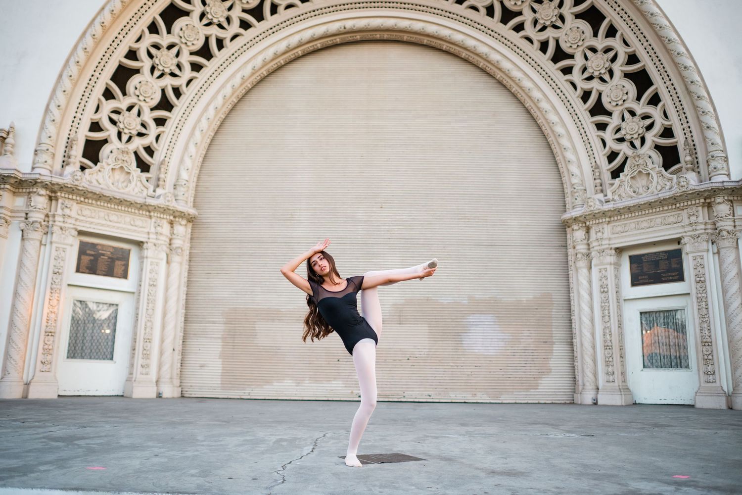 A graceful ballet dancer in a black leotard poses en pointe before an ornate arched building facade.