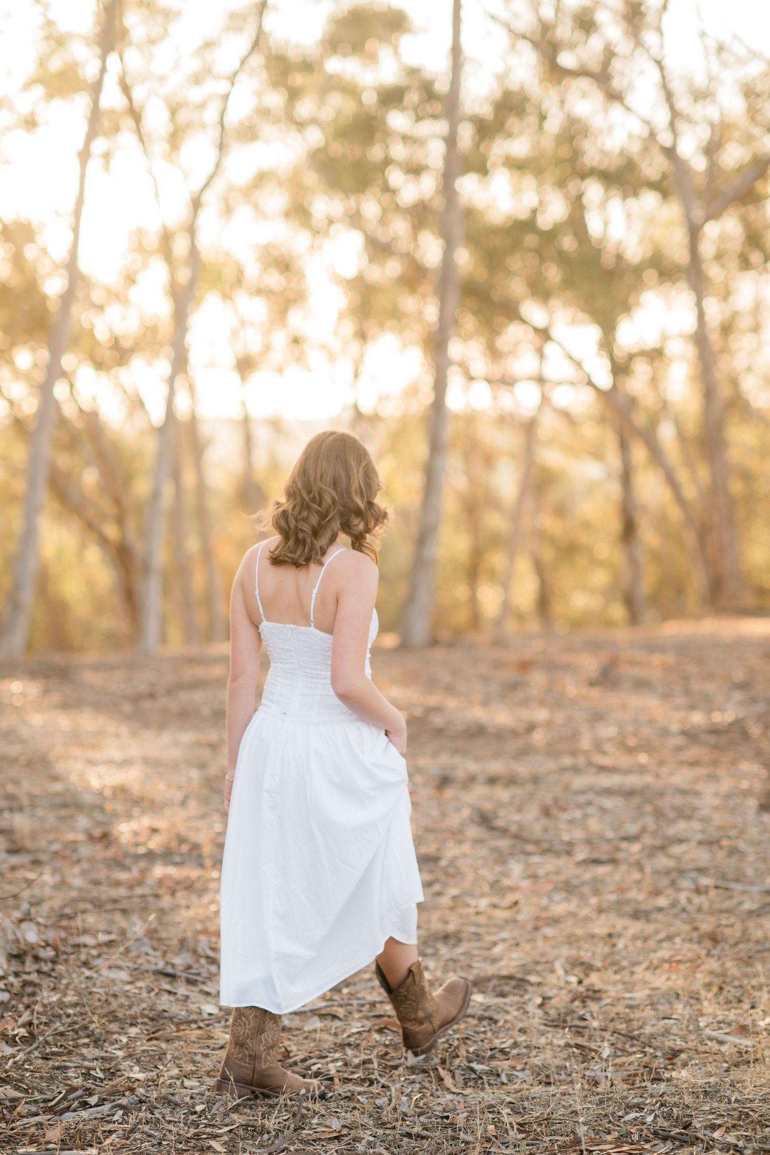 Woman in white dress and brown cowboy boots walking through golden sunlit forest, viewed from behind.