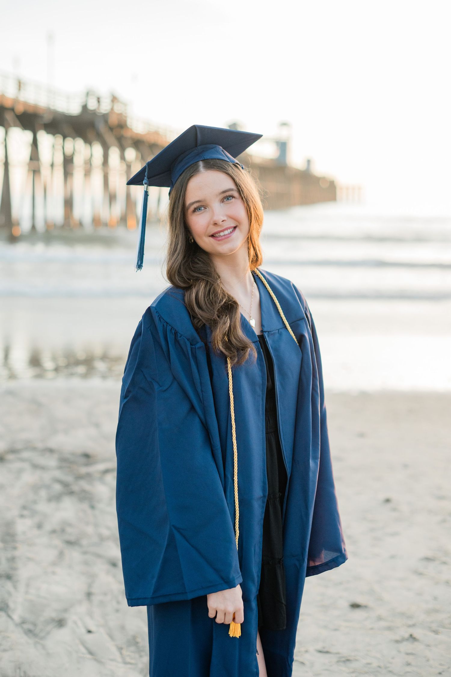 Young woman in blue graduation cap and gown smiling on a beach with a pier in the background.