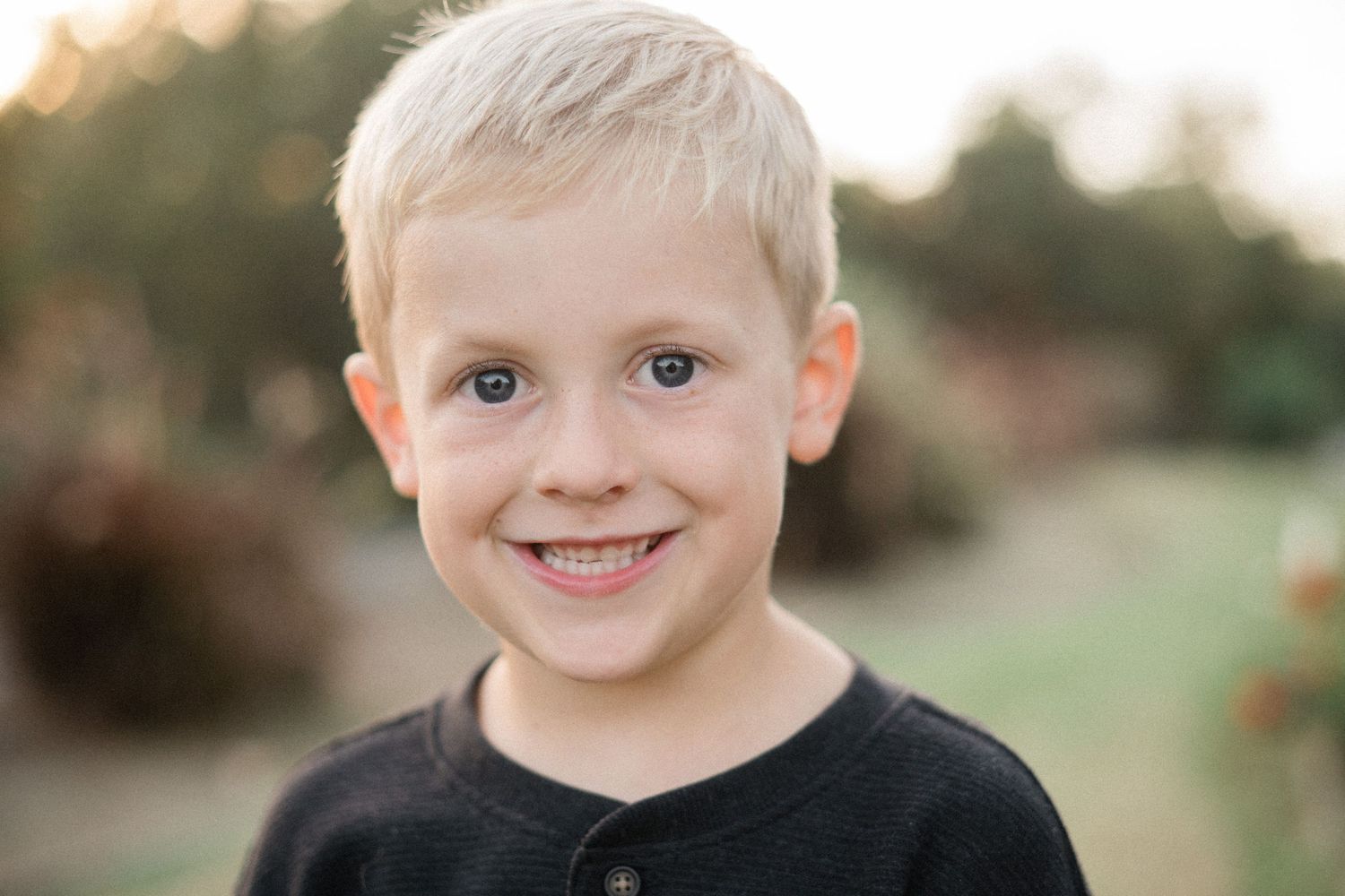 Smiling blonde boy in black shirt outdoors with blurred green background in warm natural light.