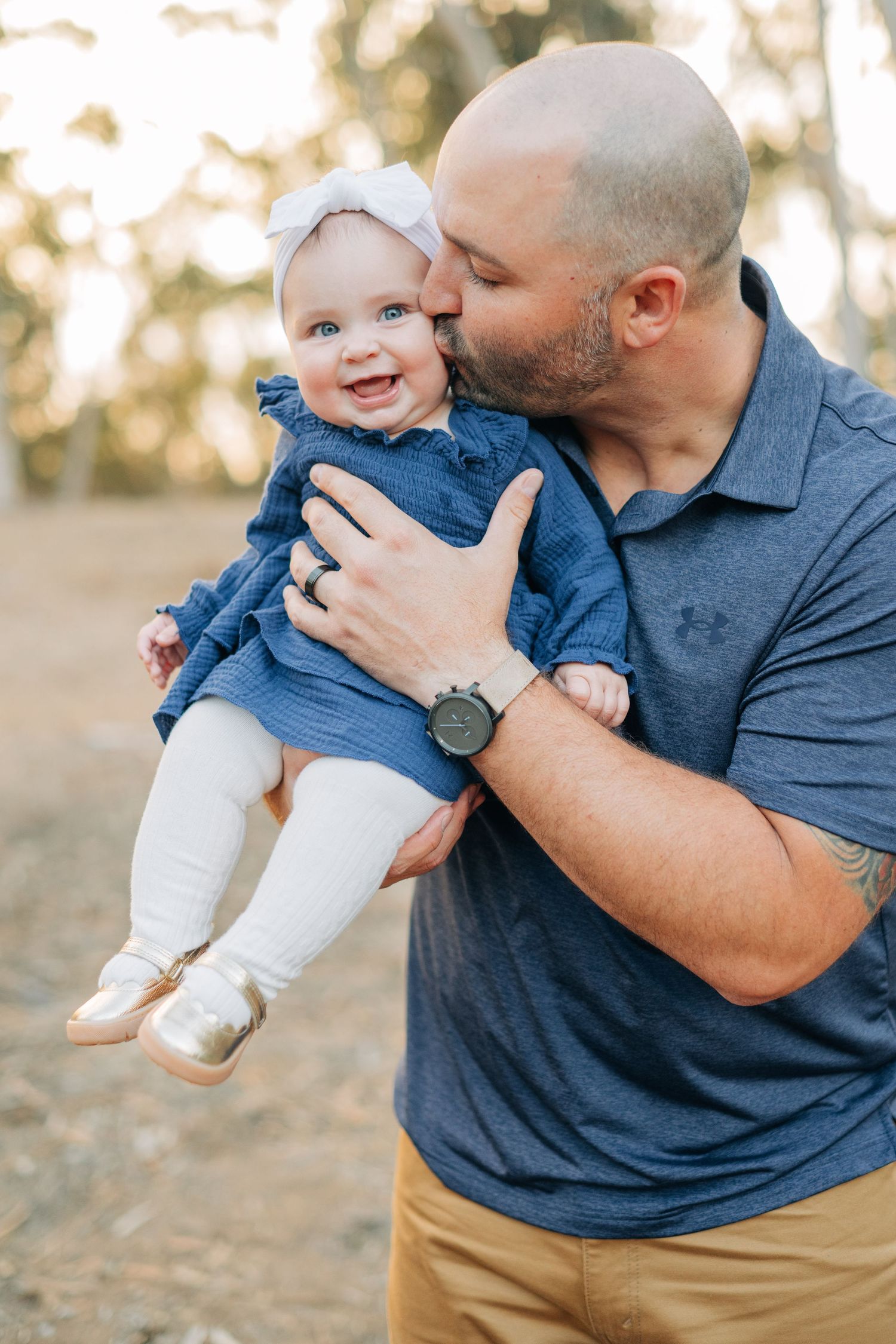Bald father kissing smiling baby girl in blue dress and white bow outdoors in golden light.