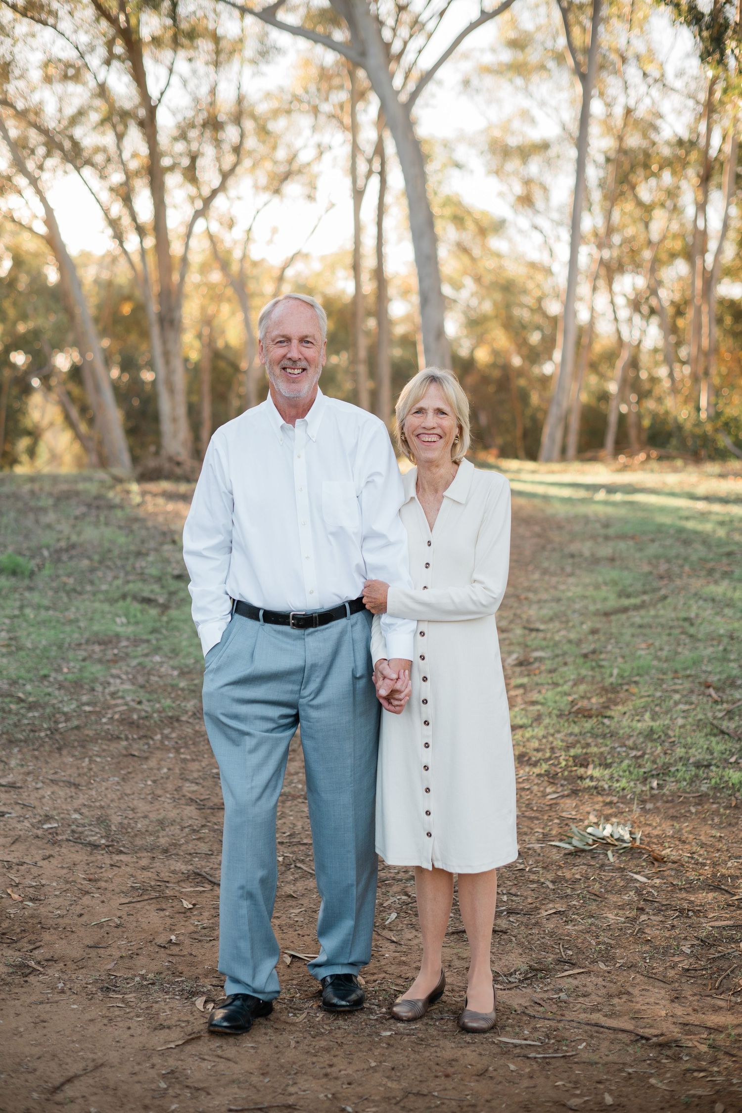 Smiling older couple standing together on a dirt path surrounded by tall eucalyptus trees in warm golden light.