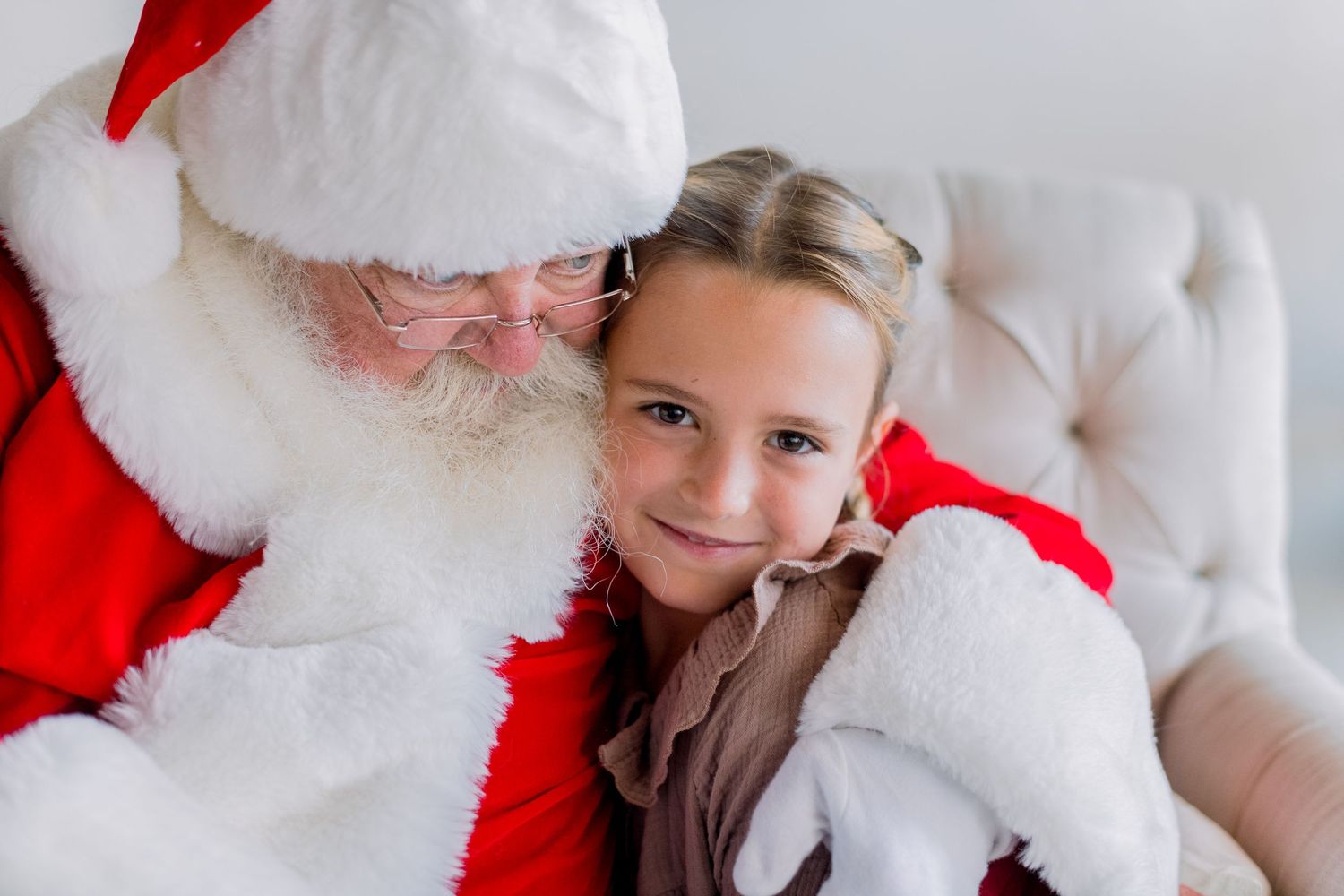 A young girl smiles while sitting with Santa Claus, both posing for a festive holiday photo together.