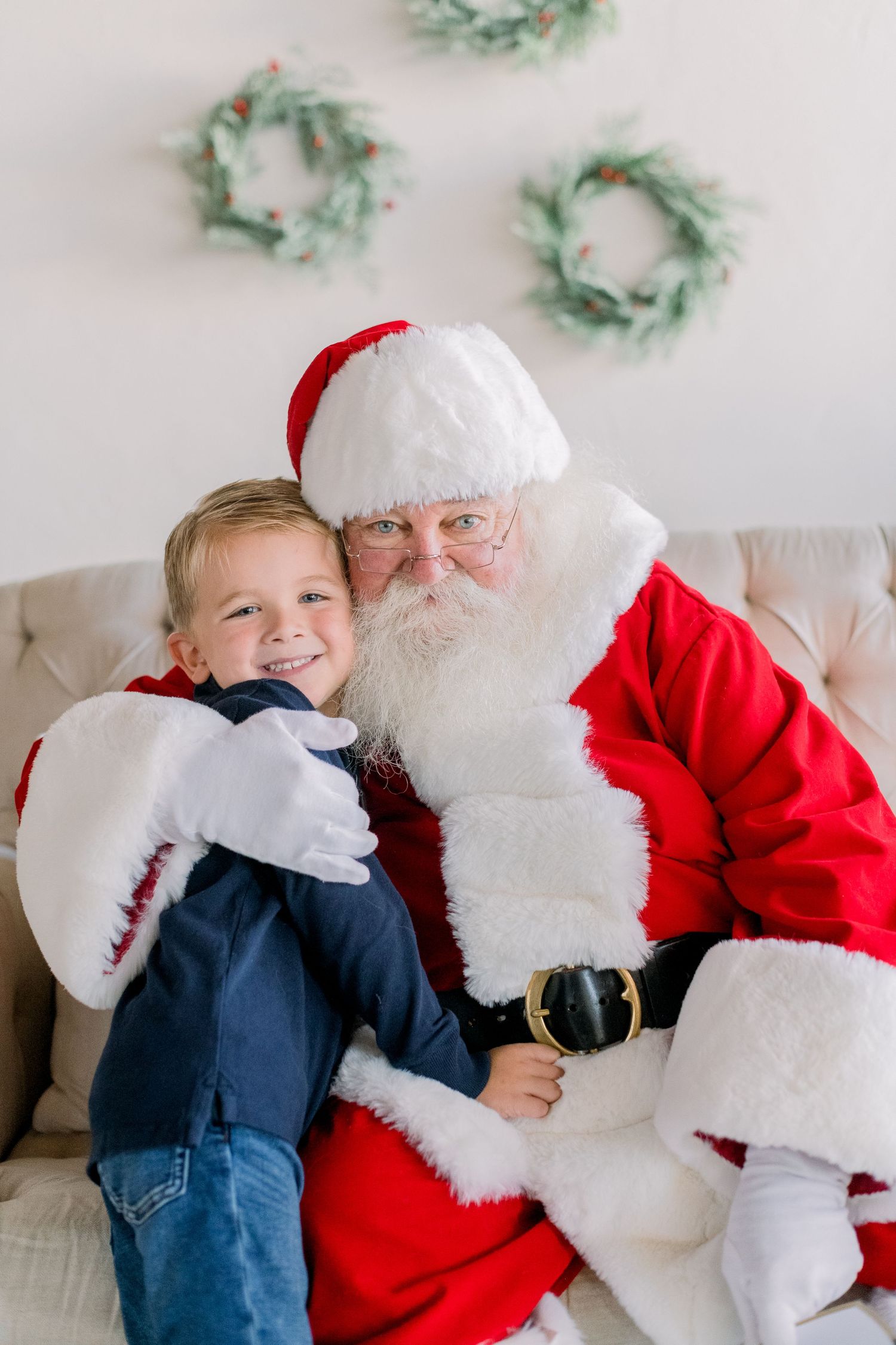 A smiling young boy sits on Santa Claus's lap in front of festive Christmas wreaths on a white wall.