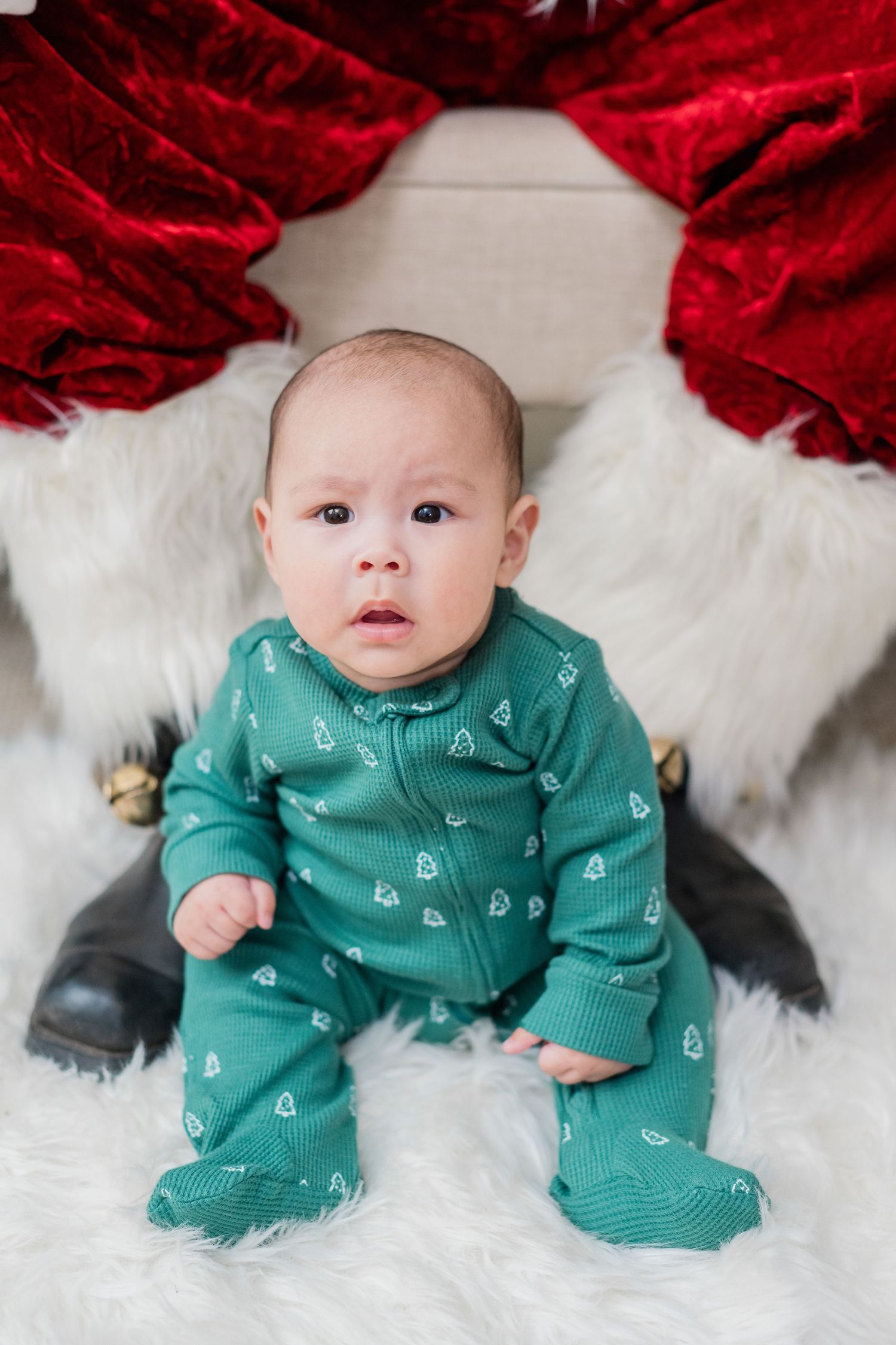Adorable baby in green Christmas tree onesie sitting on a white fur rug between Santa's legs.