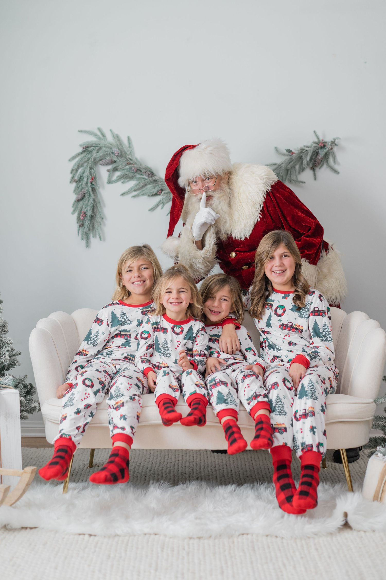 Four children in matching Christmas pajamas sit on a cream sofa with Santa Claus in a festive holiday setting.