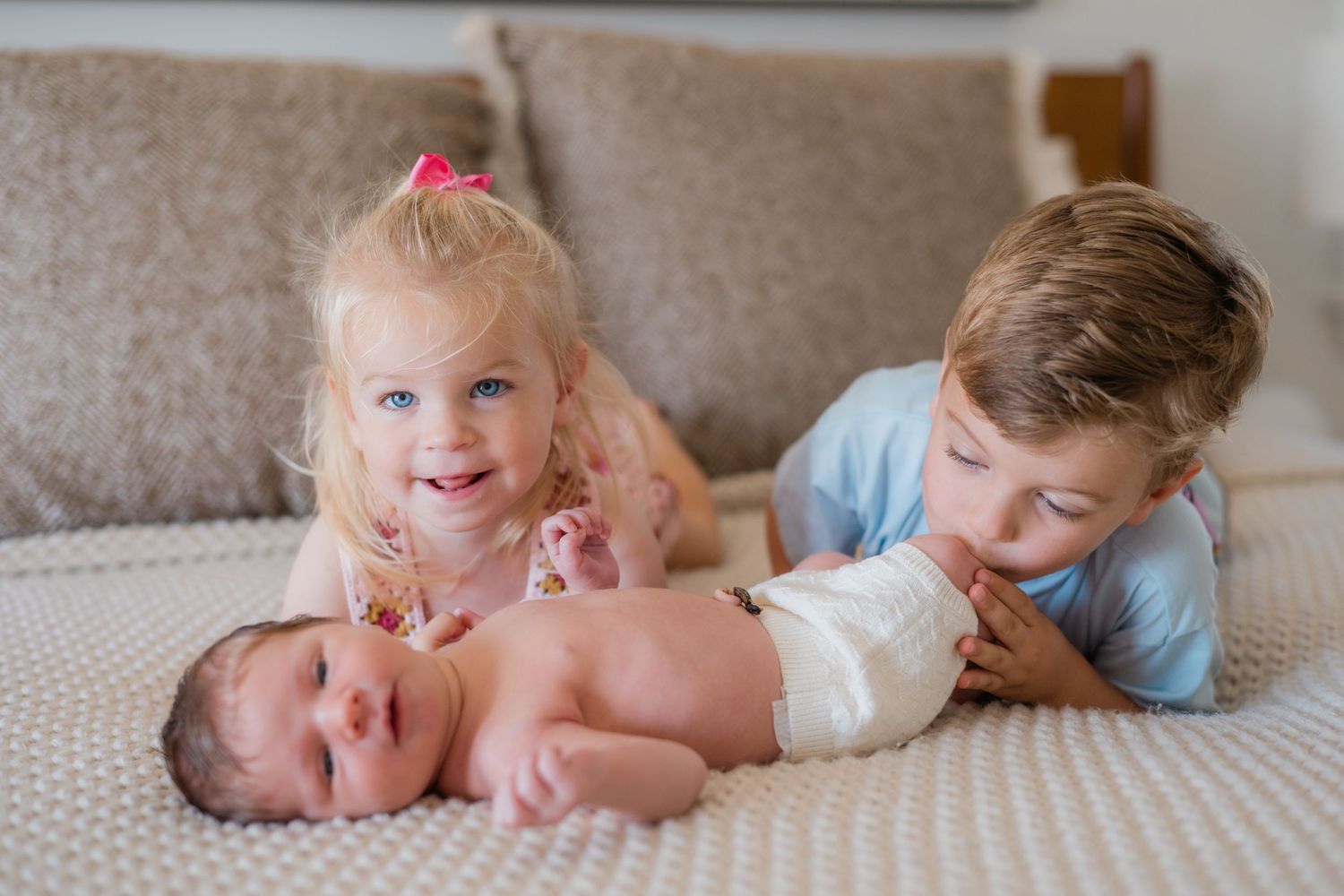 Two young siblings smile beside their newborn baby lying on a bed in a warm family portrait.