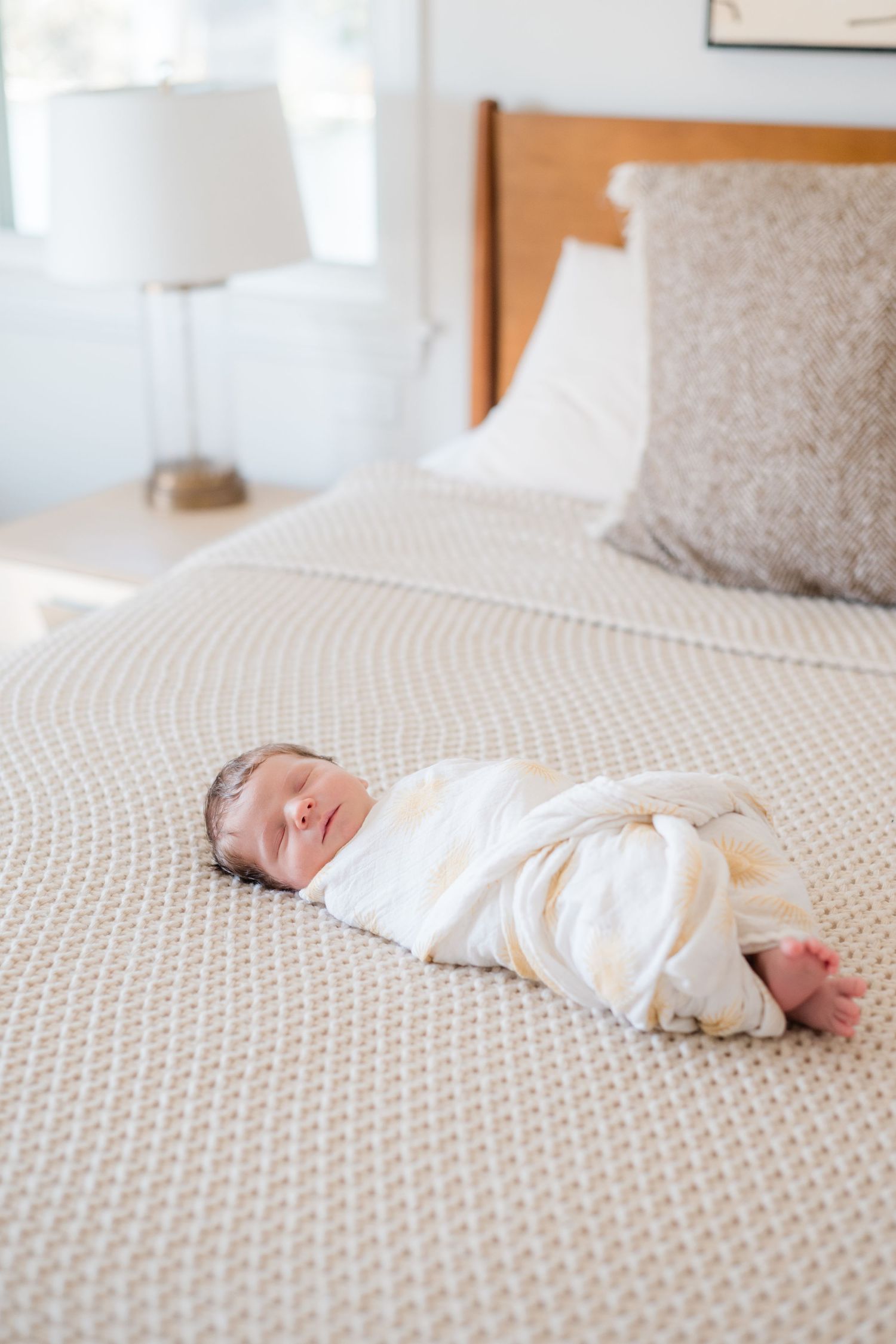 A swaddled newborn baby sleeping peacefully on a neutral textured bedspread in a bright, airy bedroom.