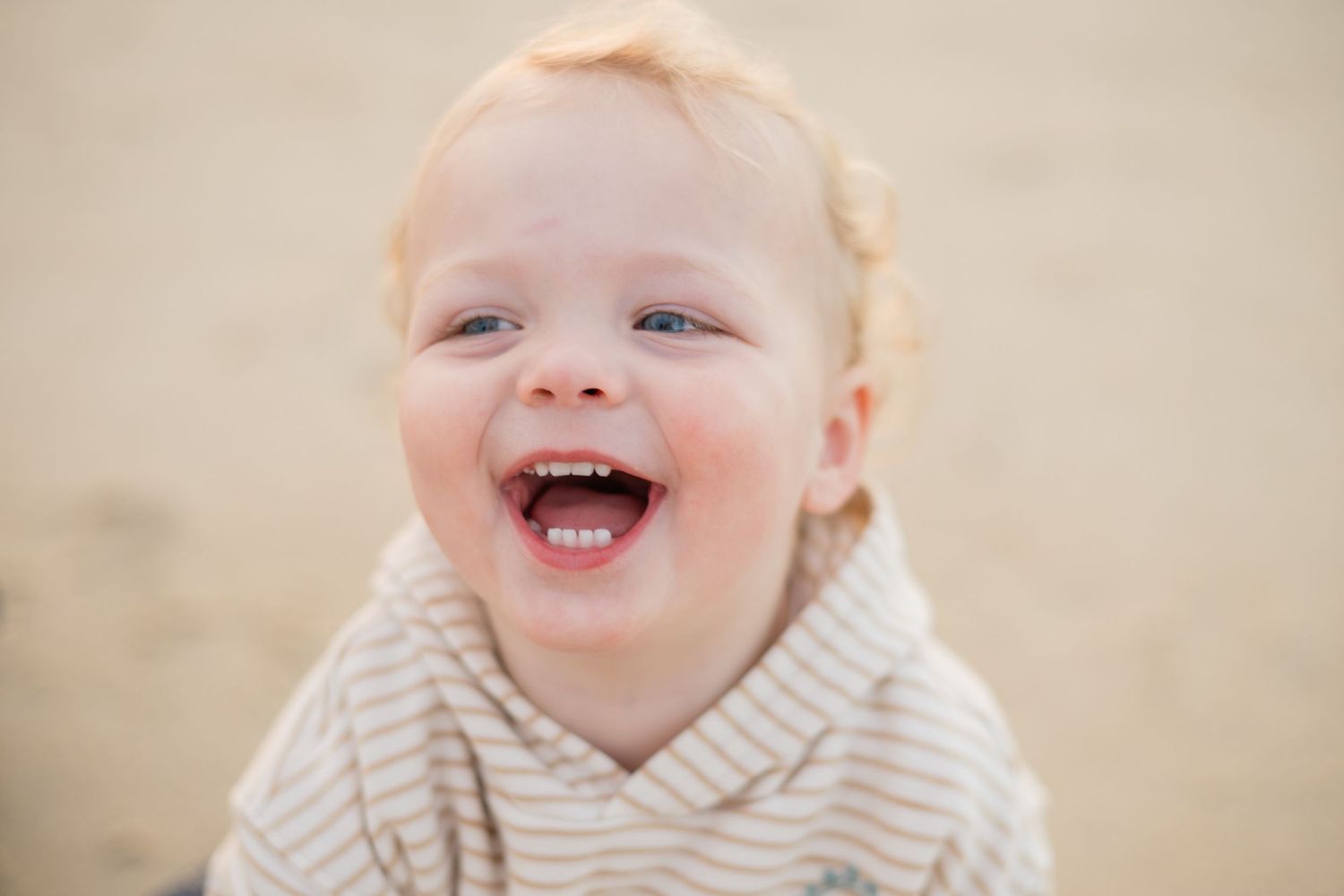 Happy laughing toddler with blonde hair and blue eyes smiling outdoors on a sandy beach.