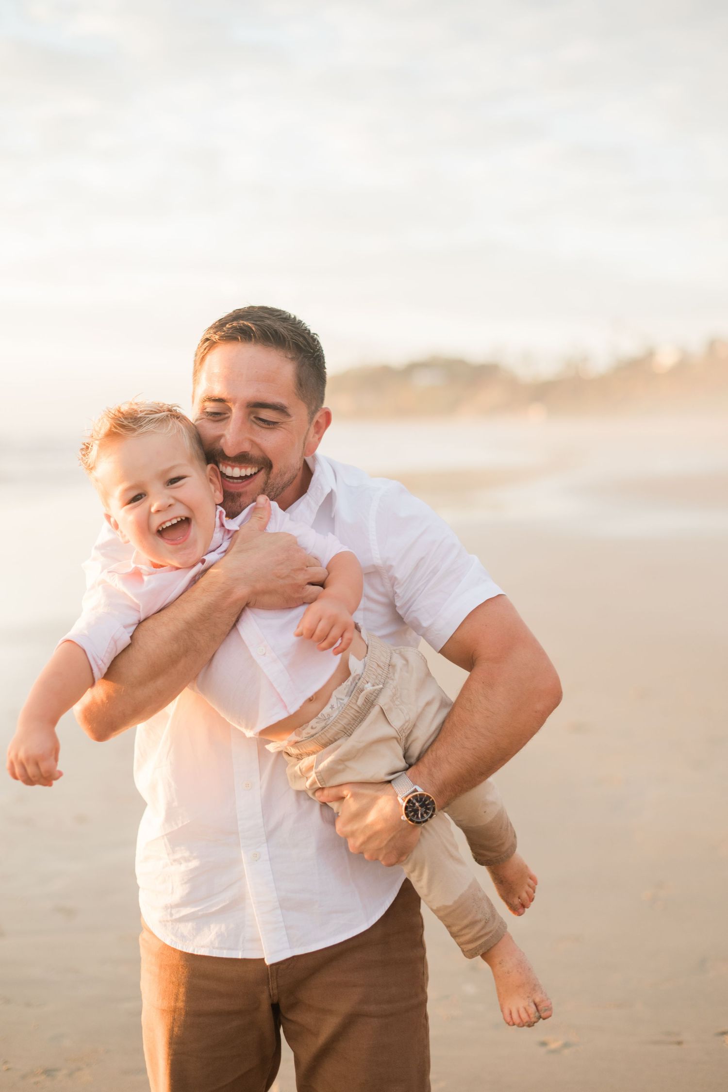 A laughing father holds his joyful toddler on a sunny beach, both wearing white shirts.